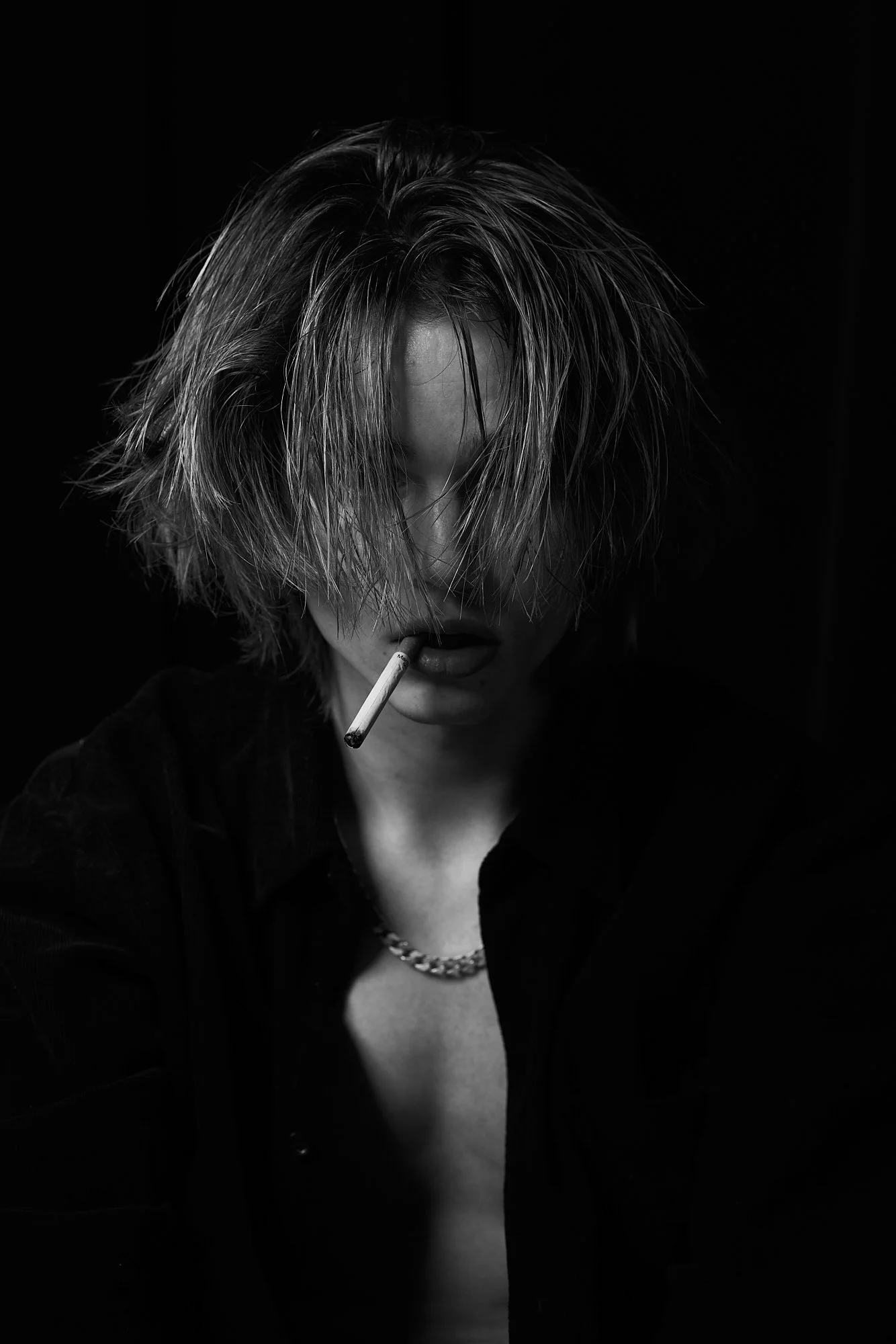 A person with messy, shoulder-length hair and a cigarette in their mouth, wearing a dark shirt and a chain necklace, in a black and white photo with dramatic lighting.
