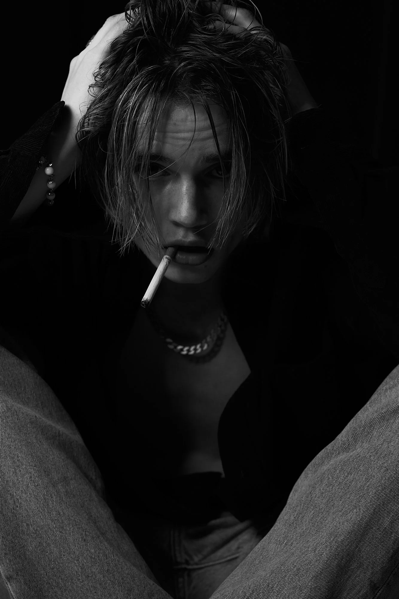 A black-and-white photo of a person with wet hair, sitting with crossed legs, holding a cigarette in their mouth, wearing a dark top, jewelry, and used to be wearing jeans.