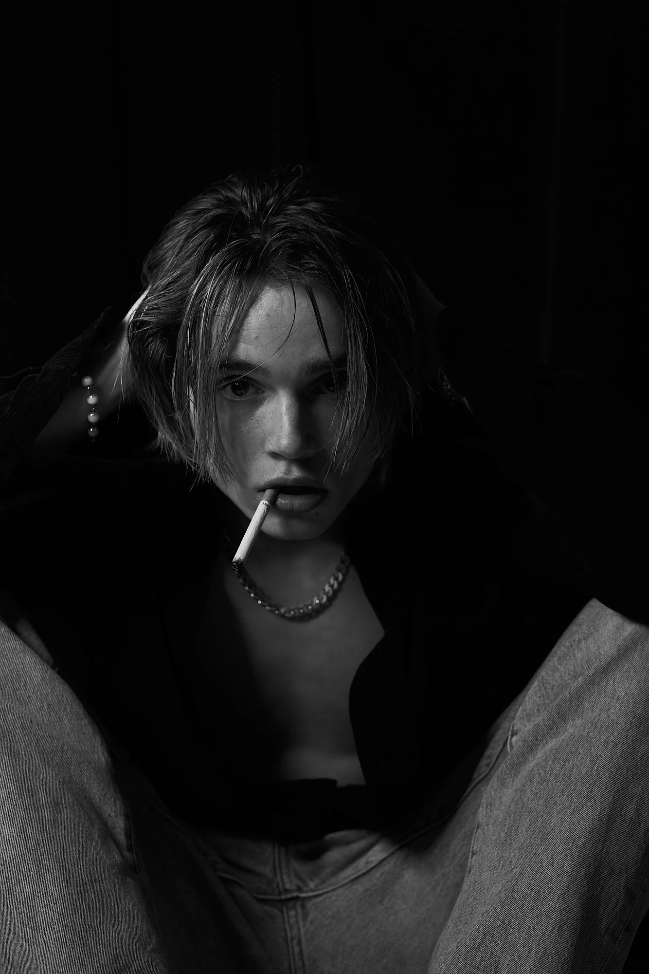A young woman with short, messy hair, wearing a necklace and bracelet, sitting with her legs apart, holding her head with both hands, and a cigarette in her mouth, in a dark, high-contrast black and white photo.