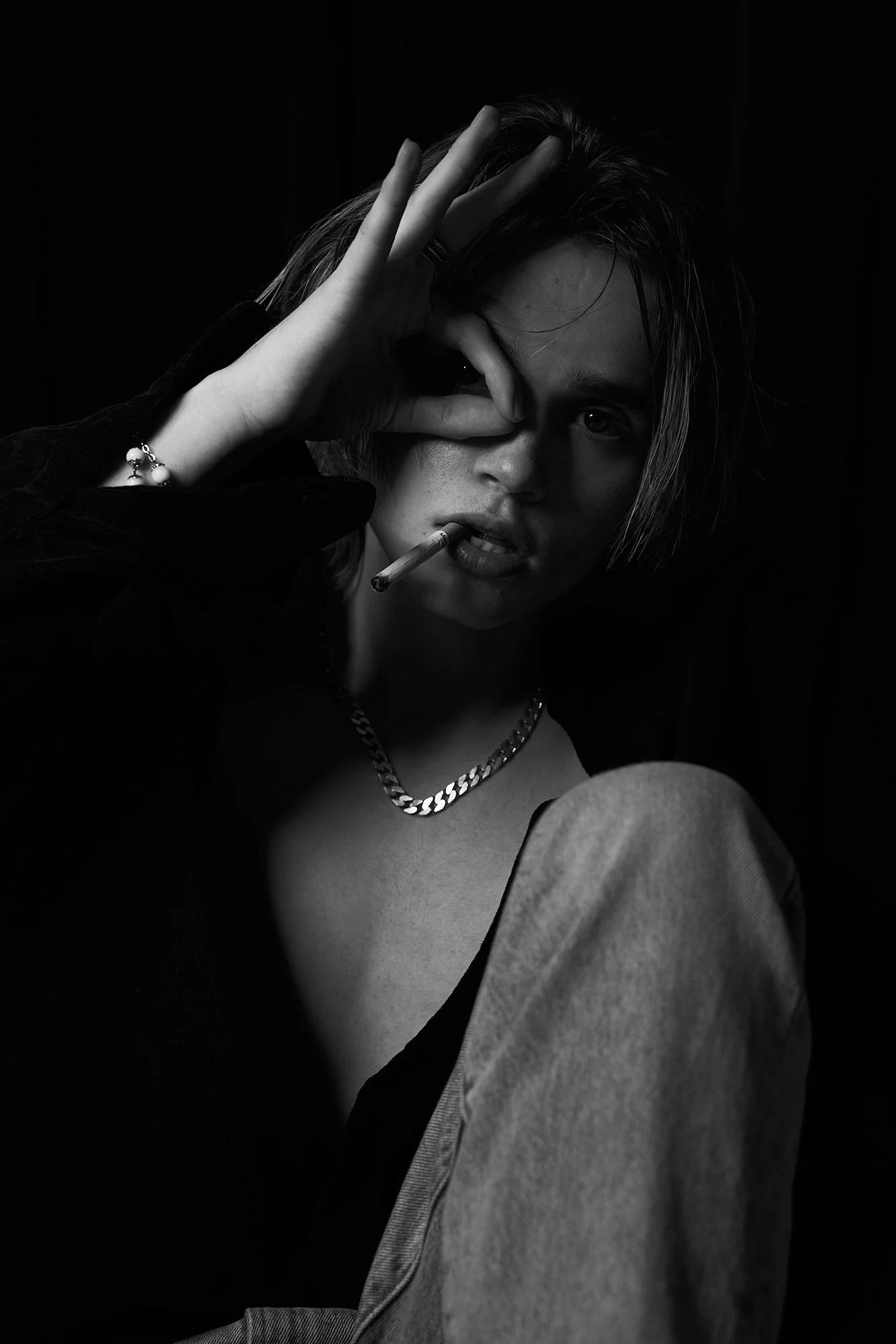 A young person with medium-length hair, wearing a chain necklace, with a cigarette in their mouth, holding one eye open with their hand, and looking directly at the camera in a black-and-white photo.