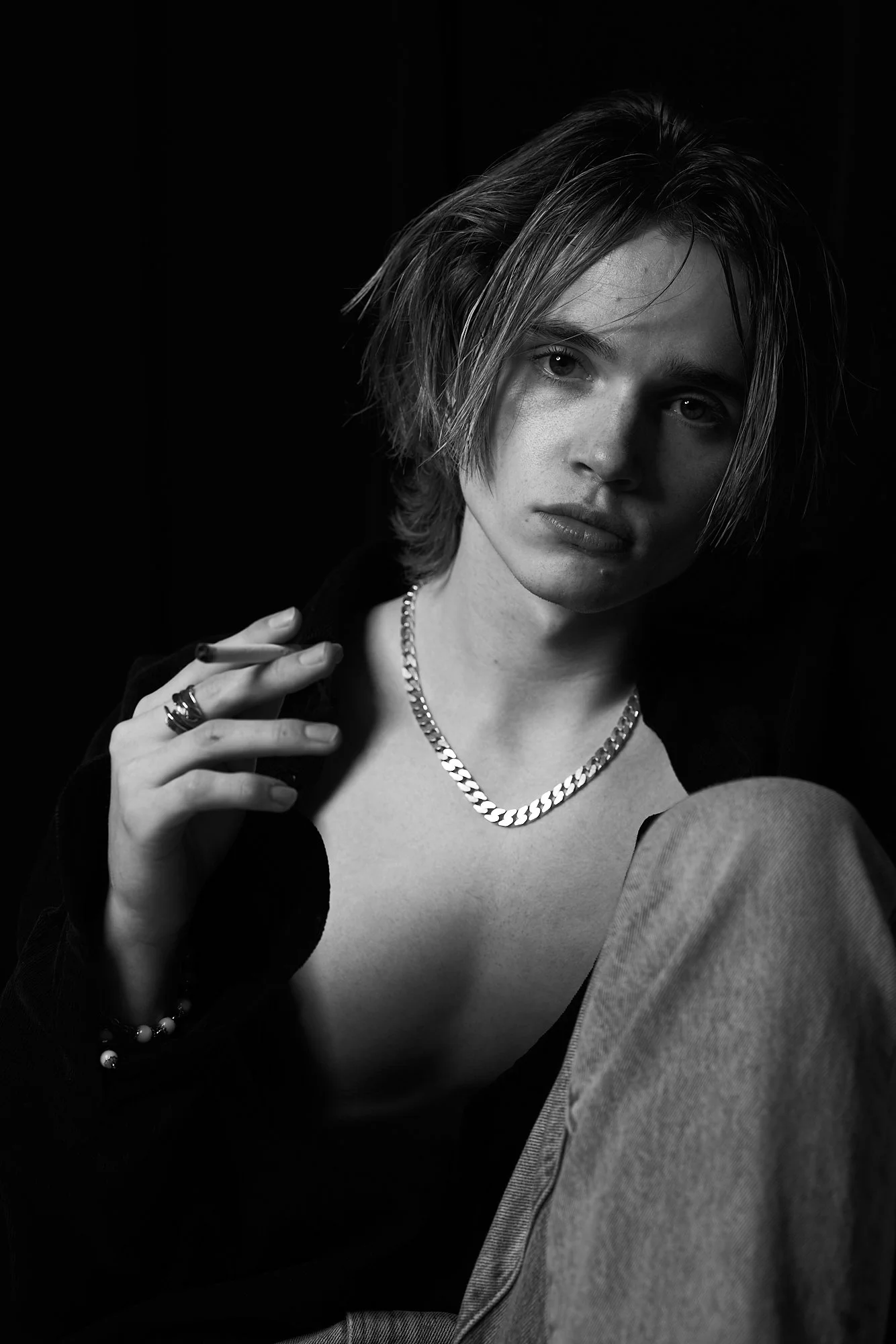 Black and white portrait of a young woman with short, tousled hair, wearing a chain necklace, rings, and a bracelet, holding a cigarette near her shoulder, with an intense expression on her face.