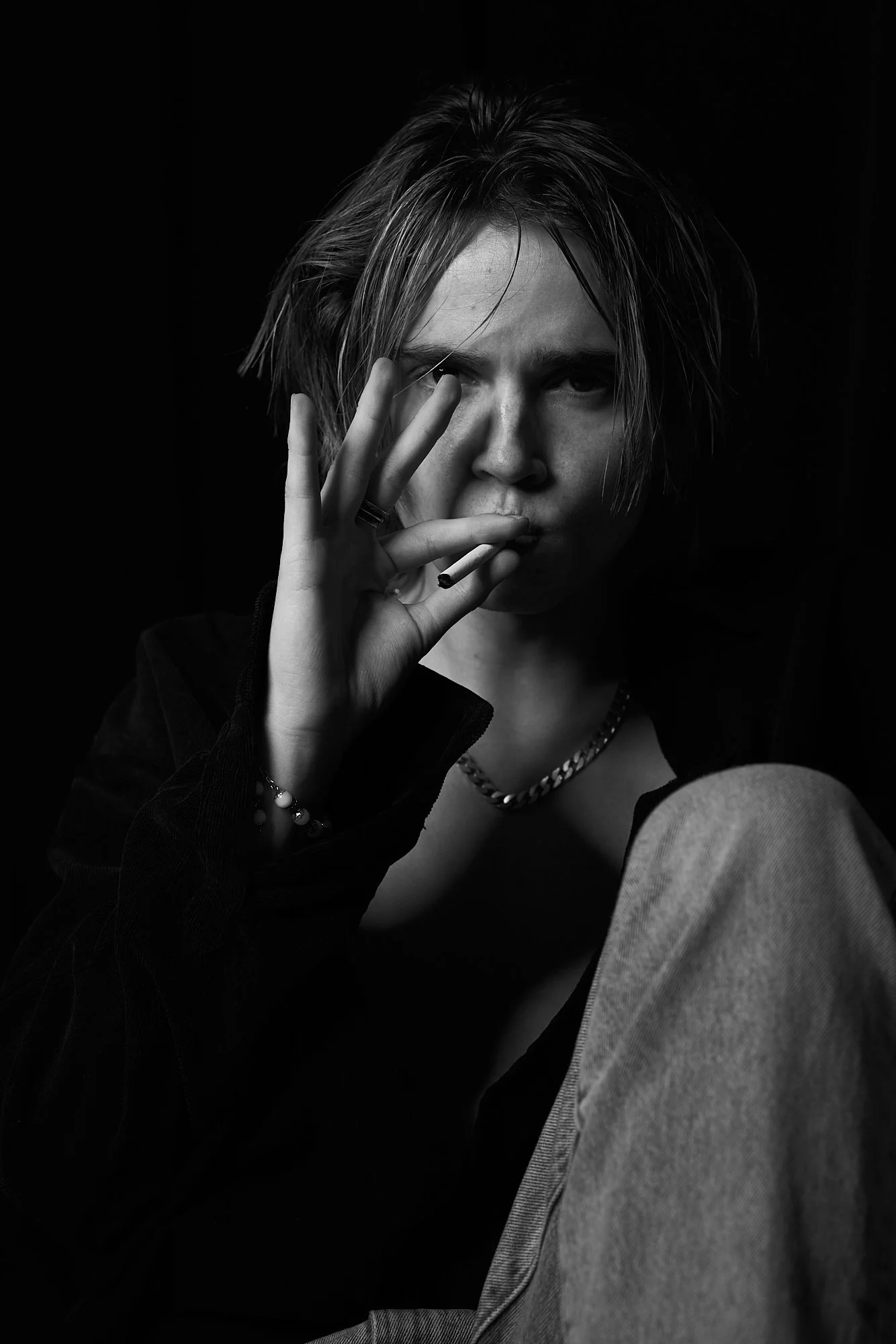 A young person with messy hair, wearing multiple rings and a chain necklace, smokes a cigarette and looks directly at the camera in a black-and-white portrait.
