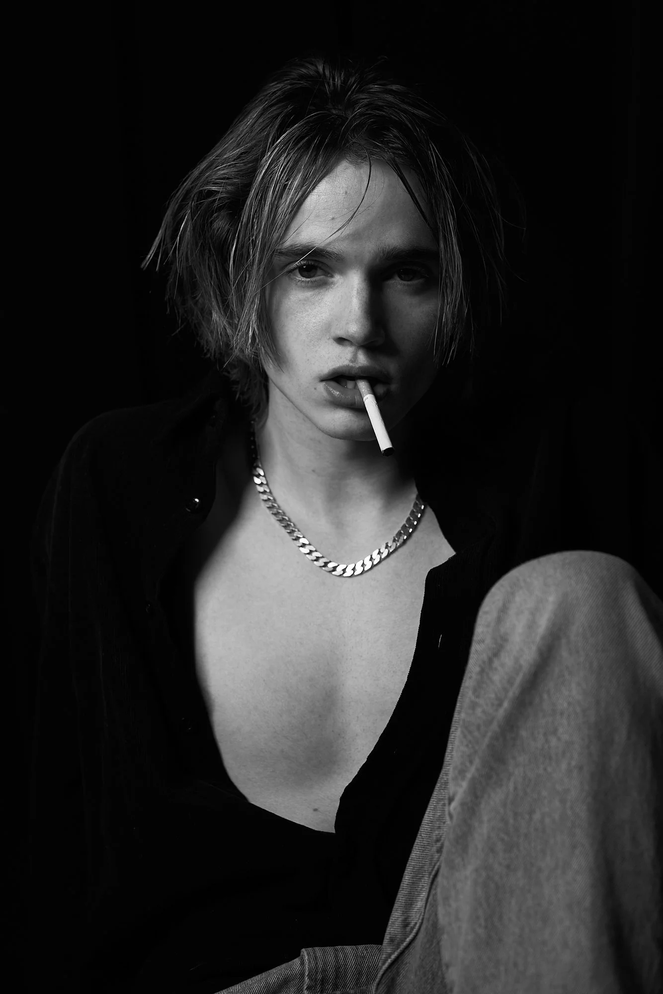 Black and white photo of a young woman with short, tousled hair, wearing a chain necklace and a dark shirt, with a cigarette in her mouth, against a dark background.
