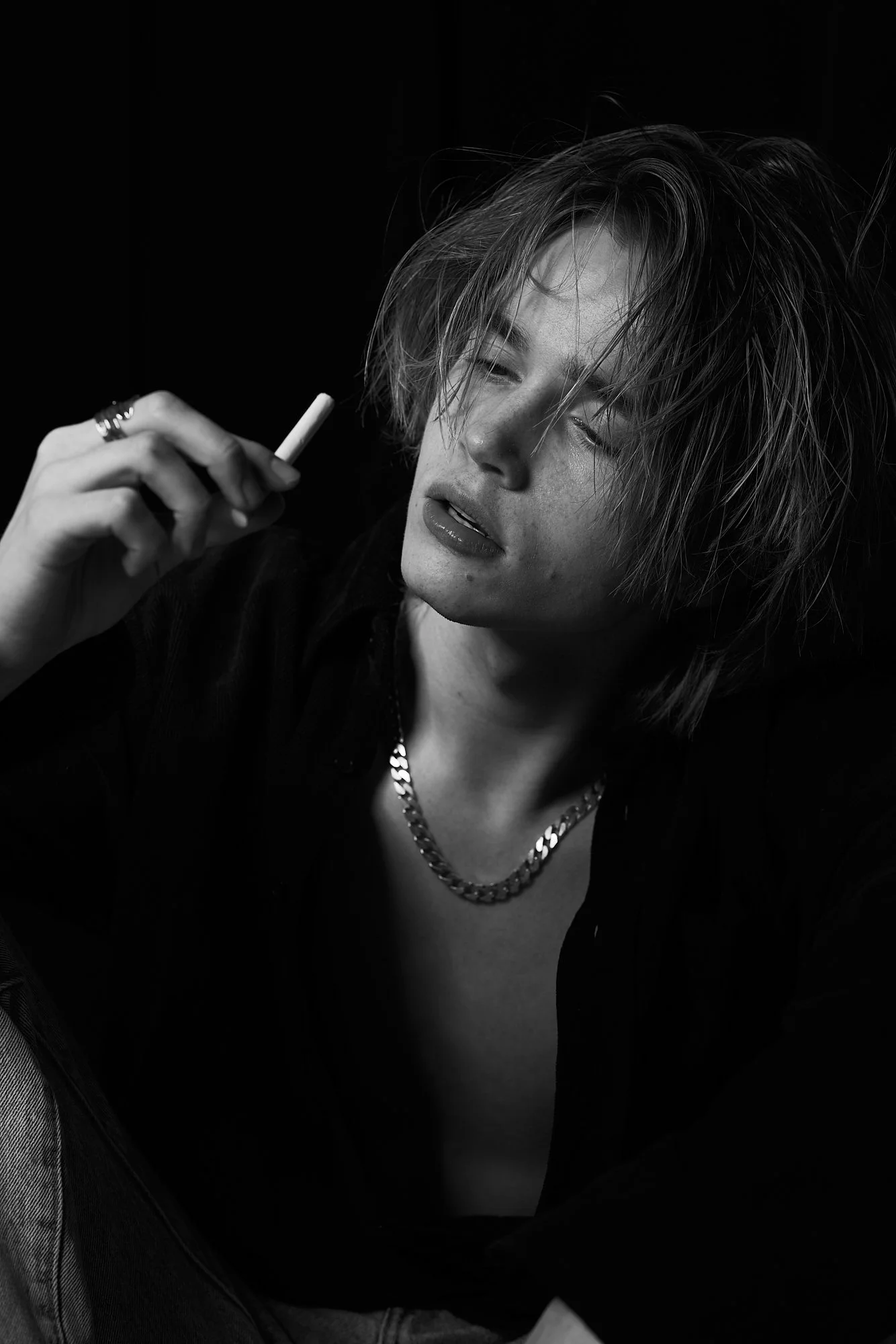 A young person with tousled hair holding a cigarette in their right hand, wearing a chain necklace and a dark shirt, with a contemplative expression, in a black and white photograph.