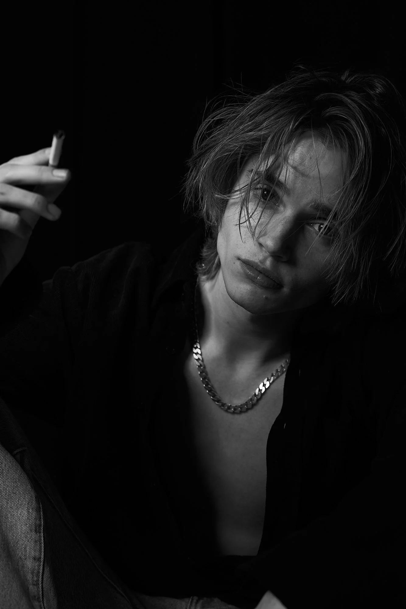 Black and white photo of a young man with messy hair, wearing a chain necklace and dark shirt, holding a cigarette, looking at the camera with a serious expression.