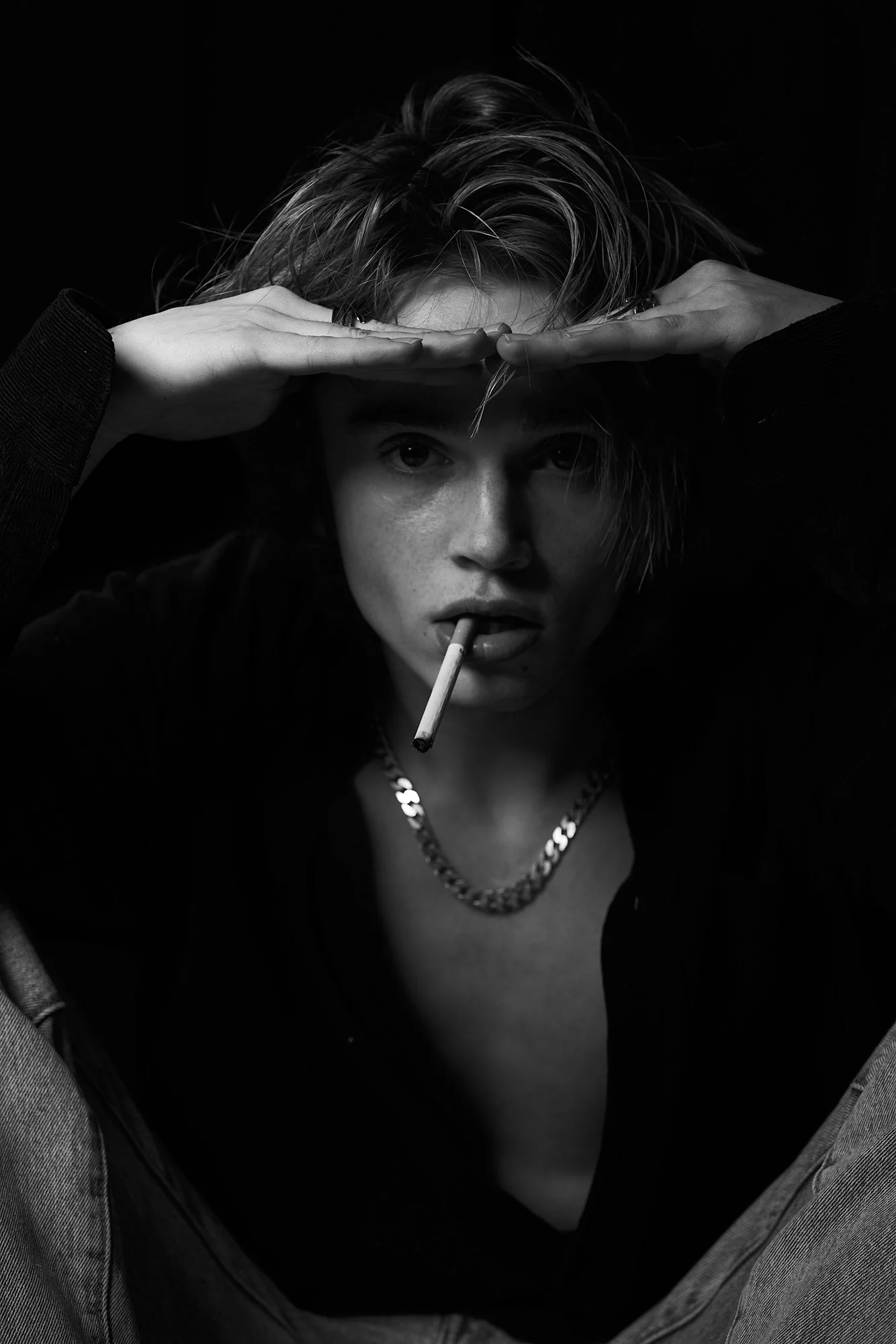 Black and white portrait of a woman with short, messy hair, looking directly at the camera with a serious expression, holding a cigarette in her mouth, and her hands pushed up on her head.