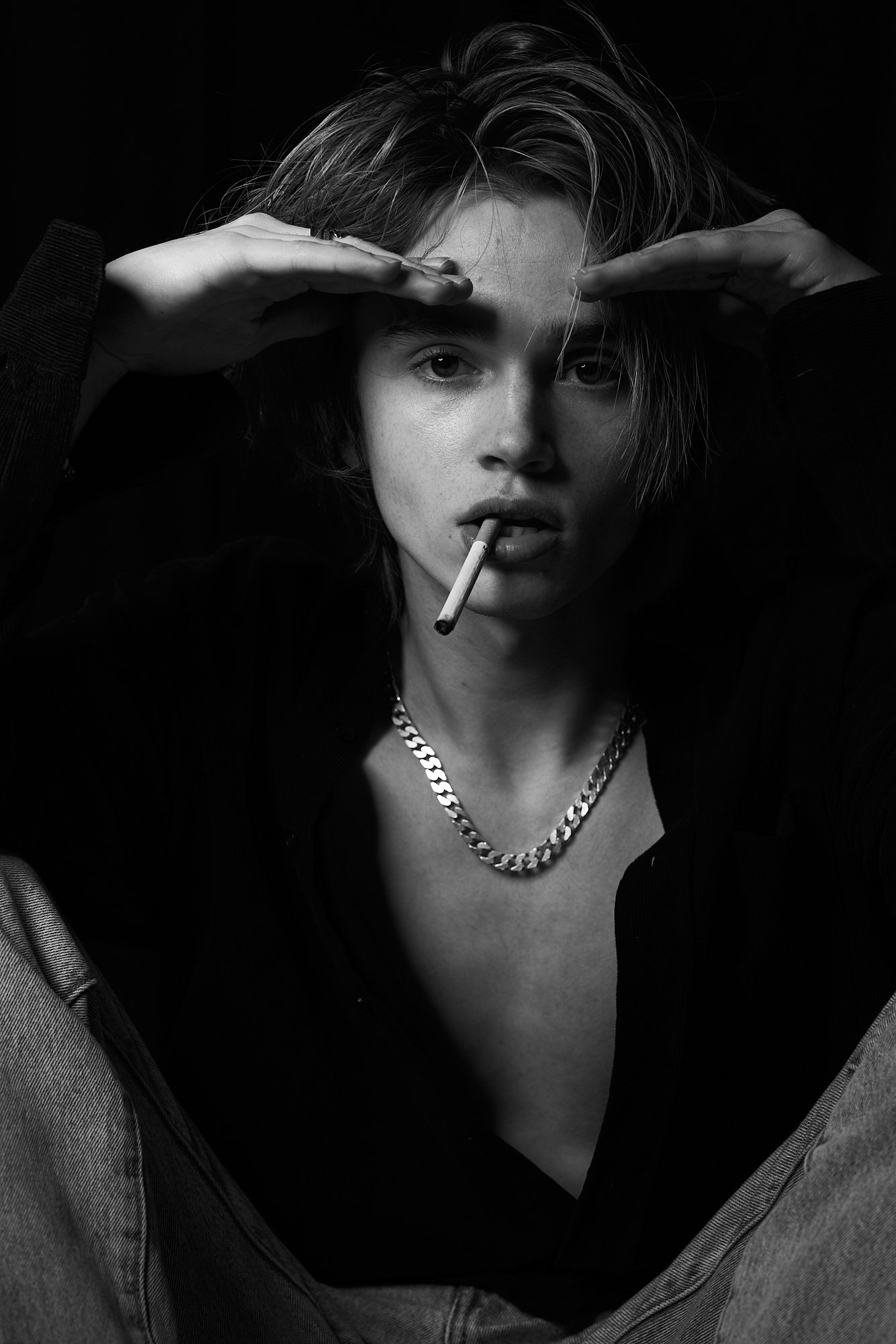 Black and white photo of a man with short hair, wearing a chain necklace, holding his hands above his head, with a cigarette in his mouth, looking directly at the camera.