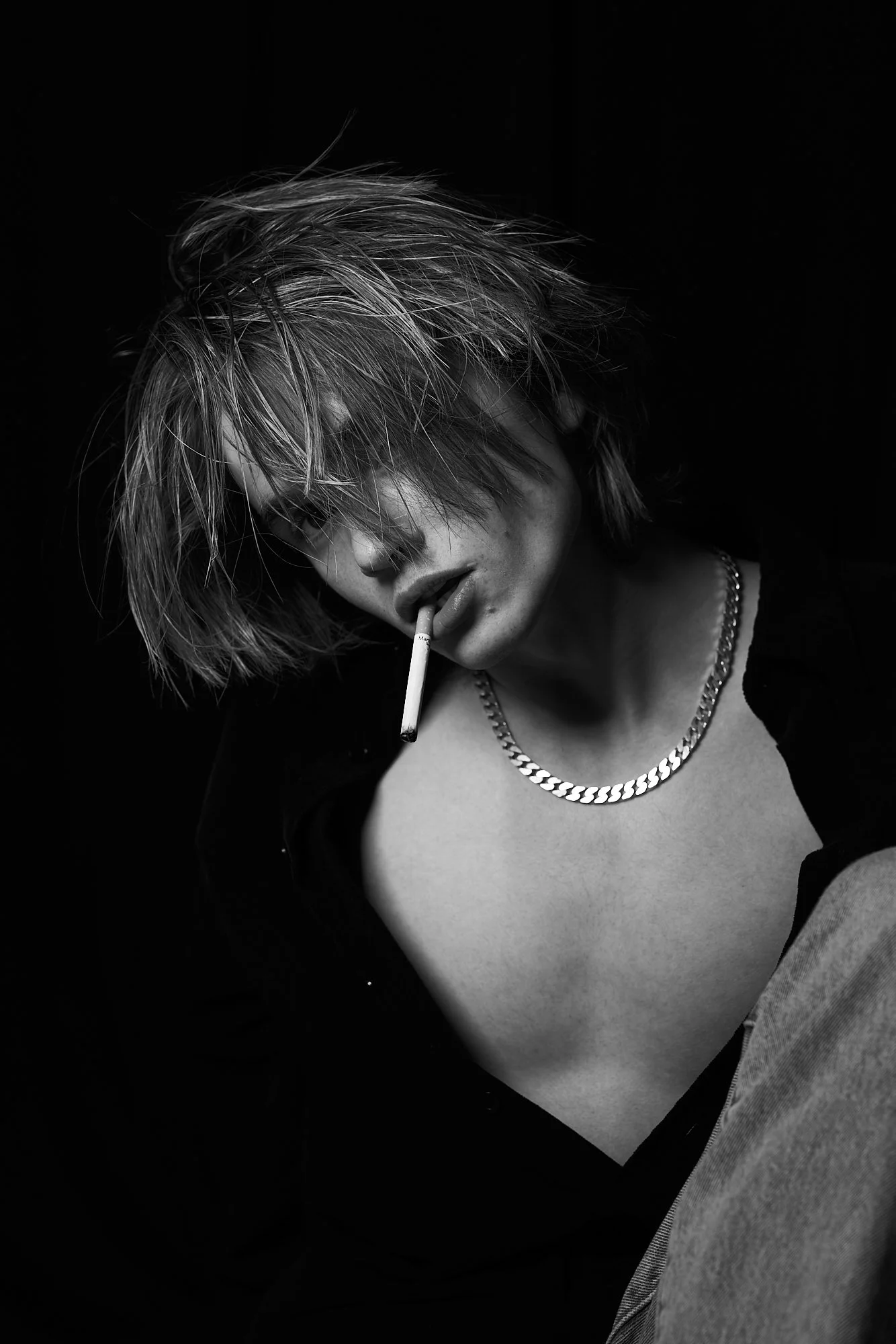 Black and white photo of a person with messy blonde hair, wearing a chain necklace and an open shirt, holding a cigarette in mouth, with a dark background.