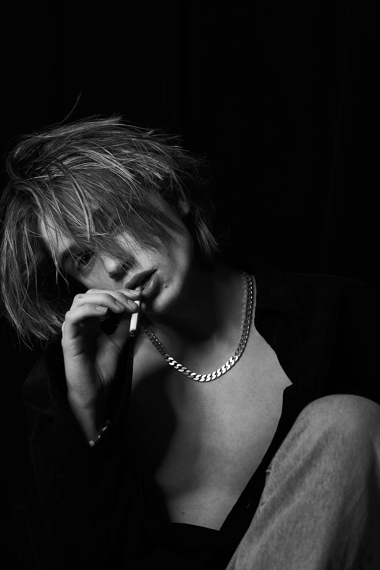 A black and white photo of a young person with messy, layered hair, wearing a chain necklace and partially unbuttoned shirt, holding a cigarette near their lips, with one eye visible and making direct eye contact with the camera.