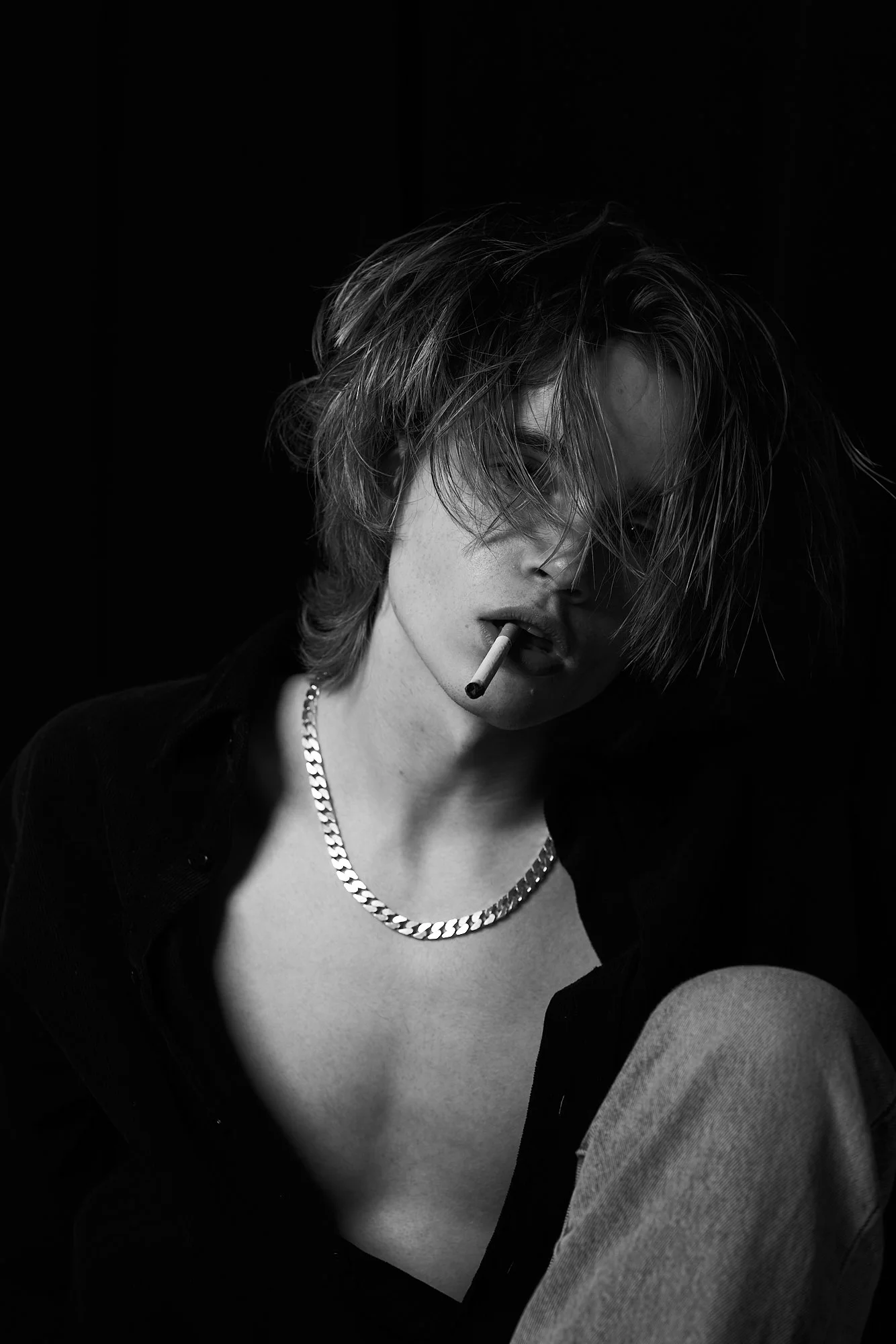 A black-and-white portrait of a young person with shaggy hair, partially covering their face, holding a cigarette in their mouth, wearing a silver chain necklace, a dark open shirt, and light-colored jeans, against a dark background.