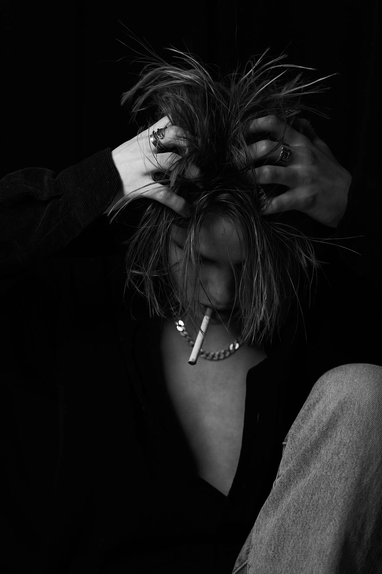 Black and white photo of a person with disheveled hair, cigarette in mouth, hands on head, wearing rings, a chain necklace, and a dark shirt.