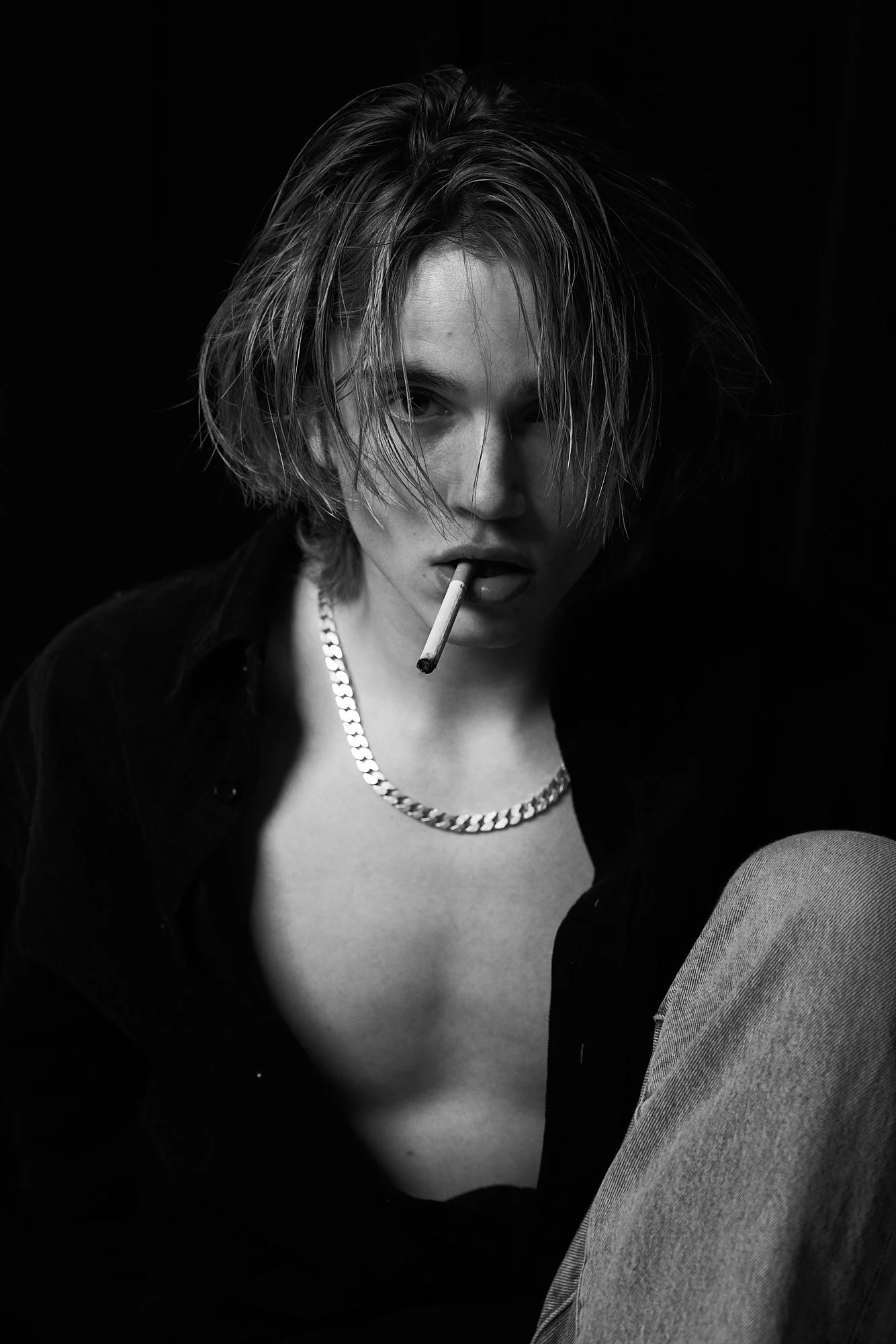 Black and white photograph of a young woman with messy short hair, wearing a chain necklace, with a cigarette in her mouth, and wearing an unbuttoned shirt revealing part of her chest, against a dark background.