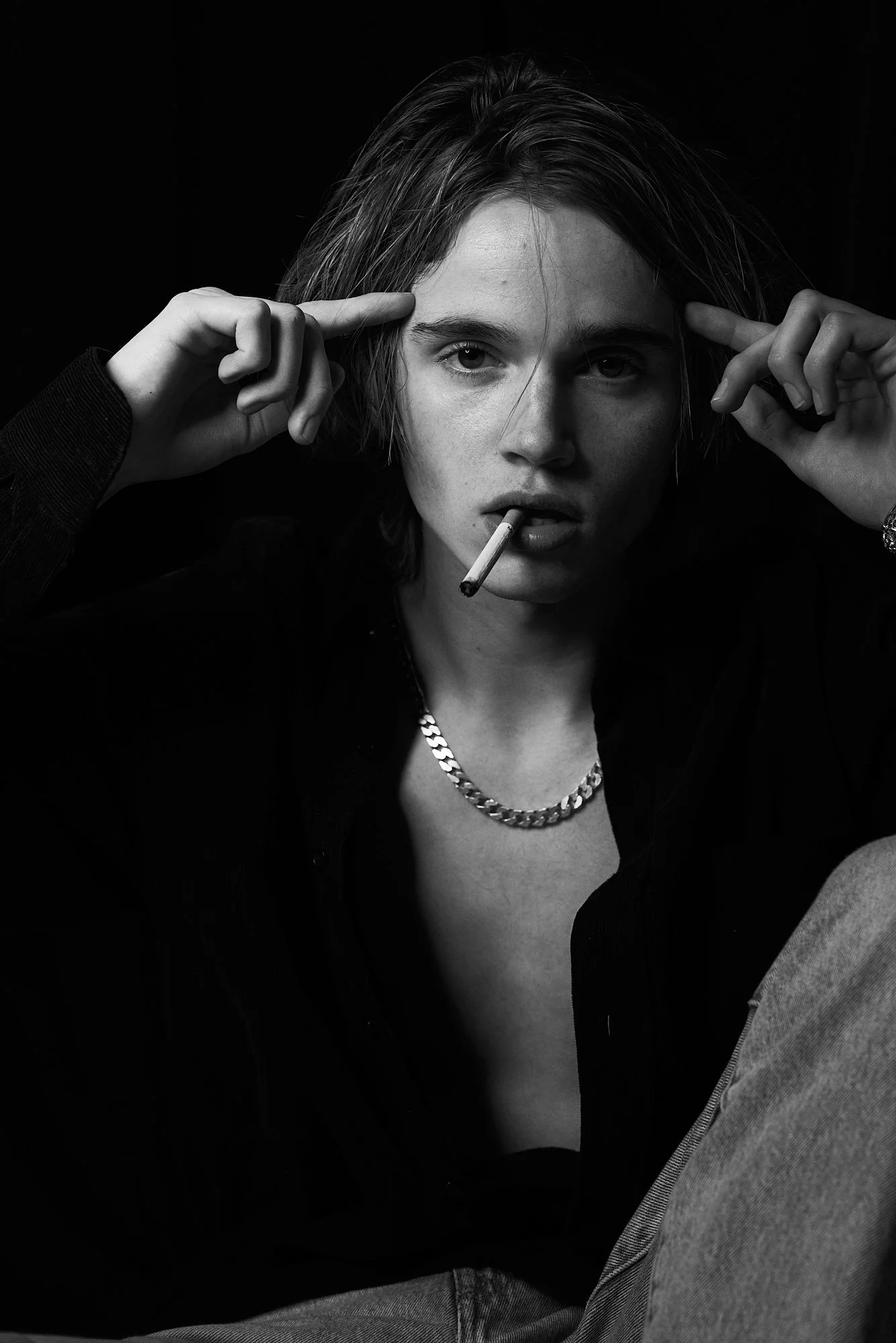Black and white portrait of a young woman with short hair, wearing a chain necklace, and a dark shirt, with a cigarette in her mouth, posing with her hands pointing to her temples.