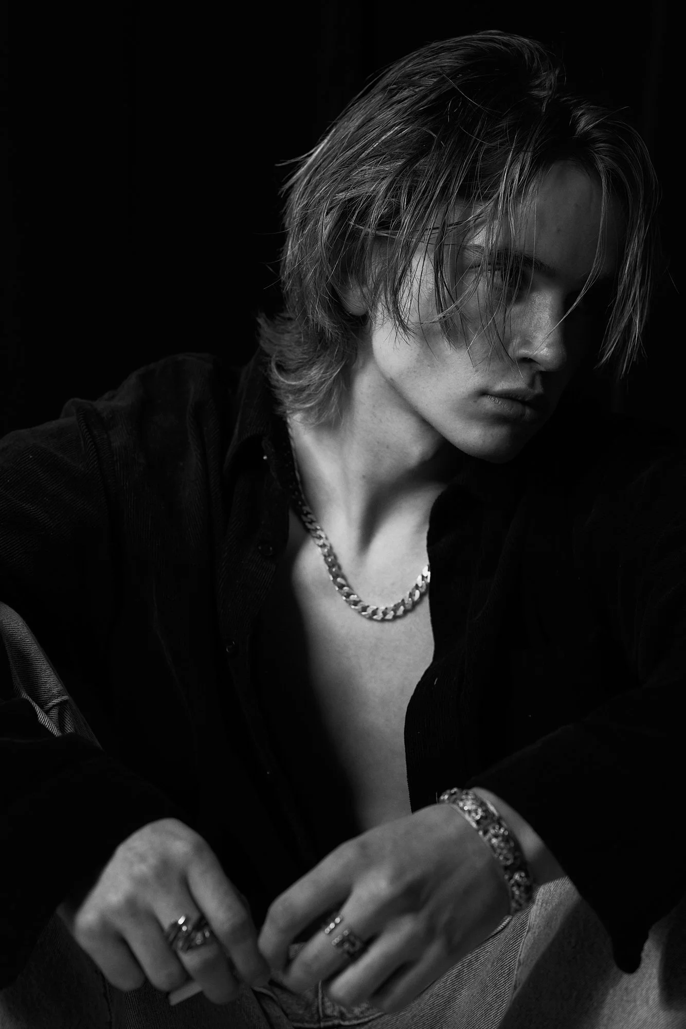 Black and white portrait of a young man with shoulder-length hair, wearing multiple rings, a chain necklace, a bracelet, and a dark shirt, posing with a serious expression.
