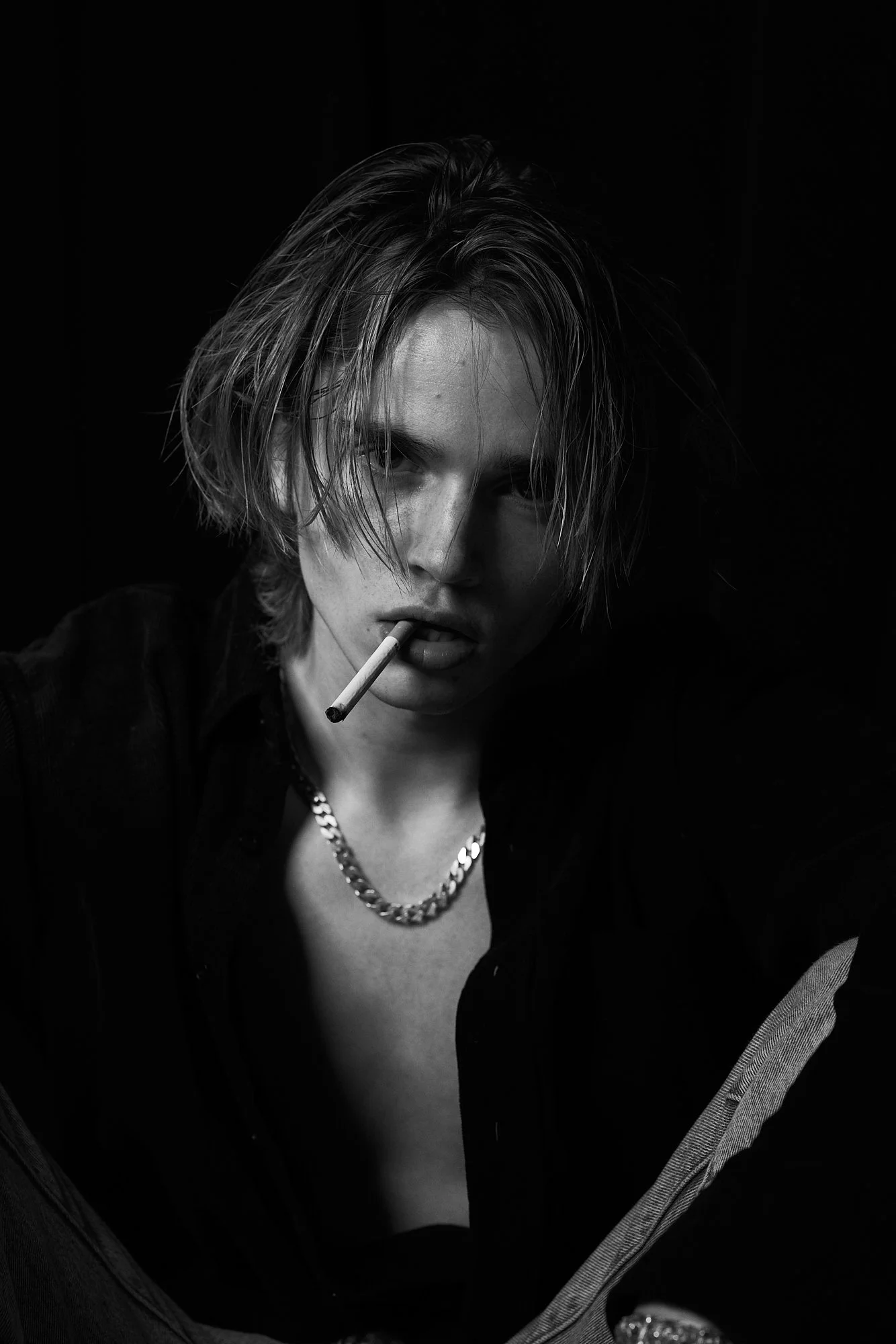 Black and white portrait of a young man with messy medium-length hair, wearing a chain necklace, with a cigarette in his mouth, looking directly at the camera in a dark background.