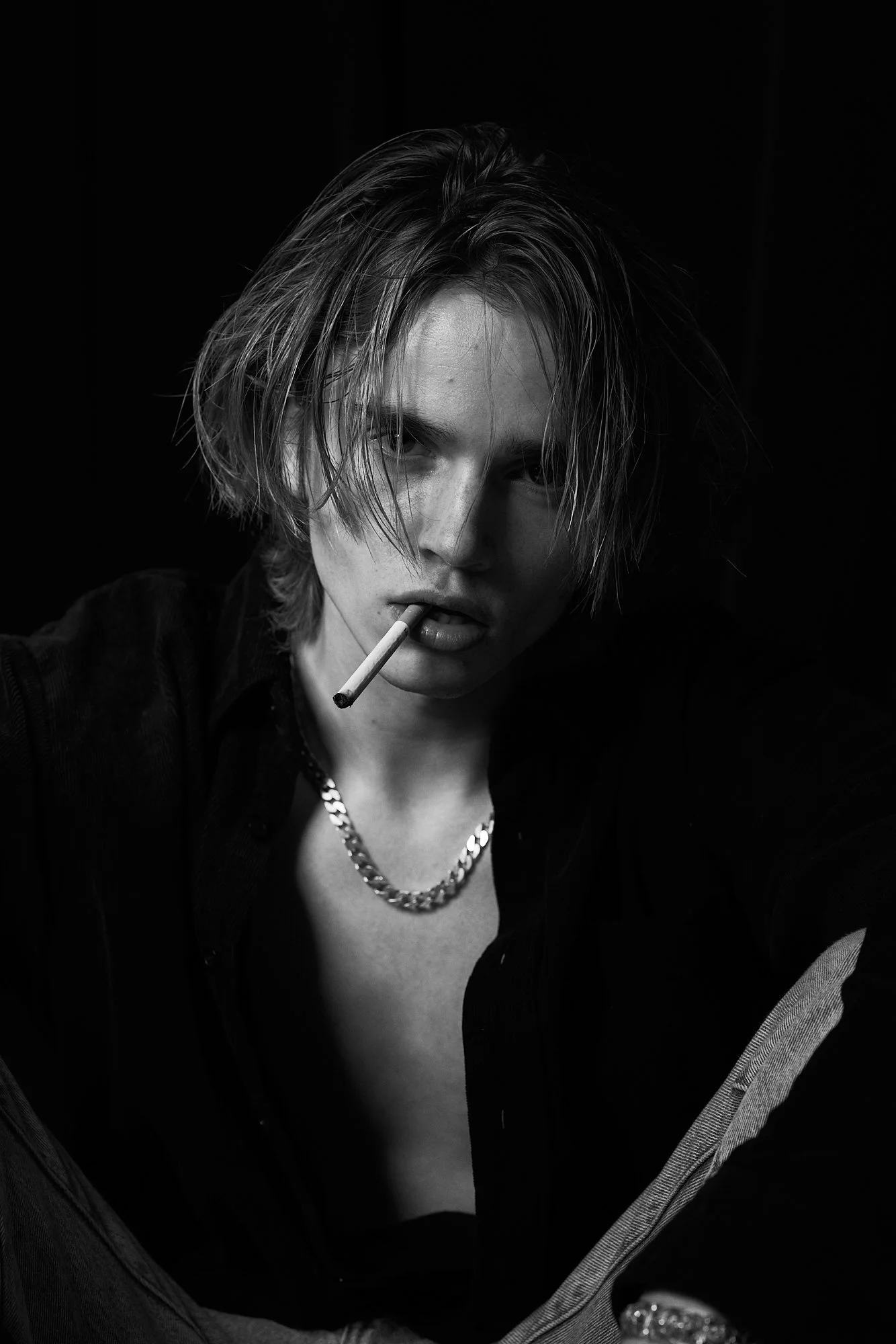 Black and white portrait of a young person with medium-length, messy hair, wearing a chain necklace, black shirt, with a cigarette in their mouth, looking intensely at the camera.