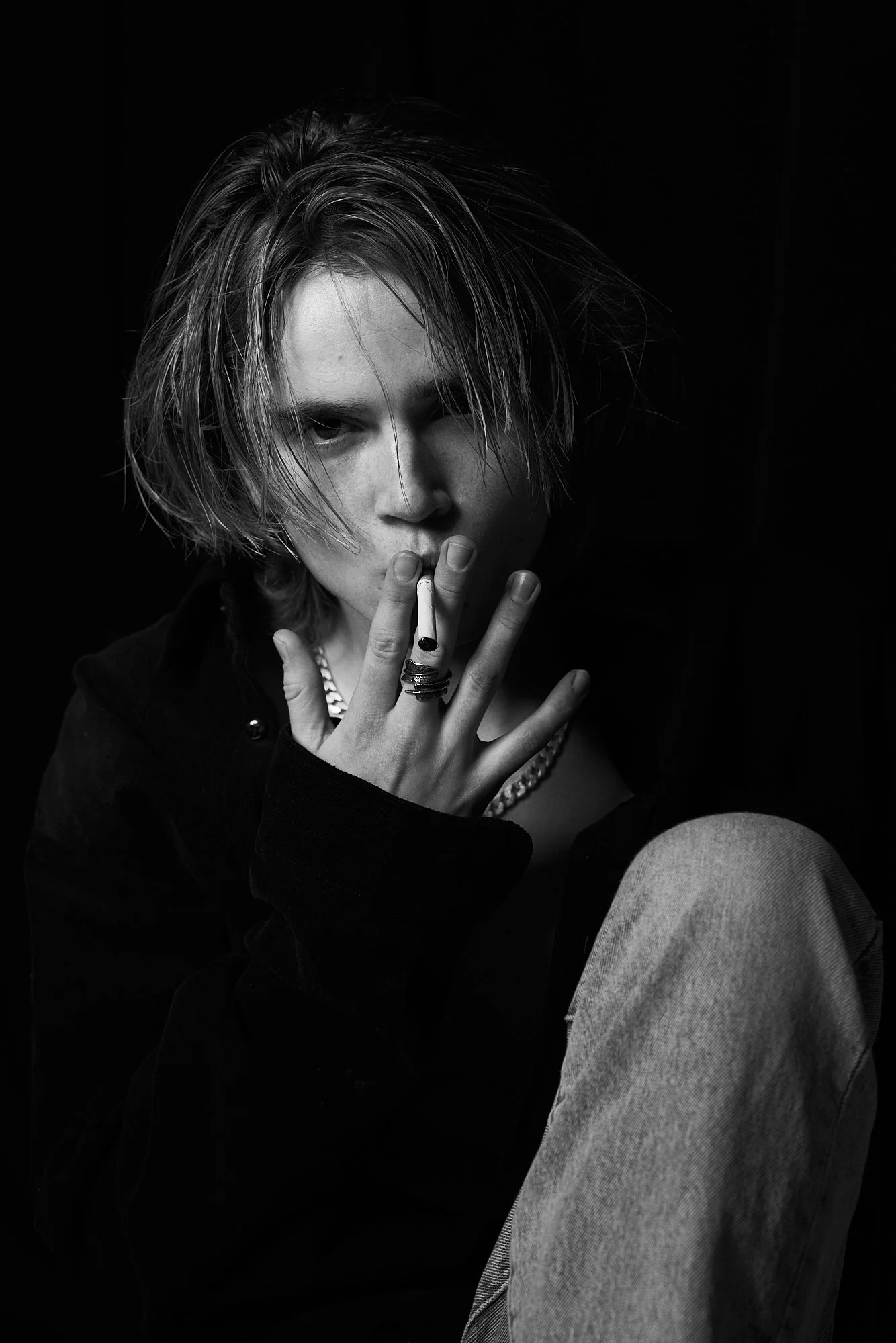 A black and white portrait of a young person with wavy, shoulder-length hair, partially covering one eye, smoking a cigarette, wearing rings, a chain necklace, a dark jacket, and light-colored pants, with a dark background.