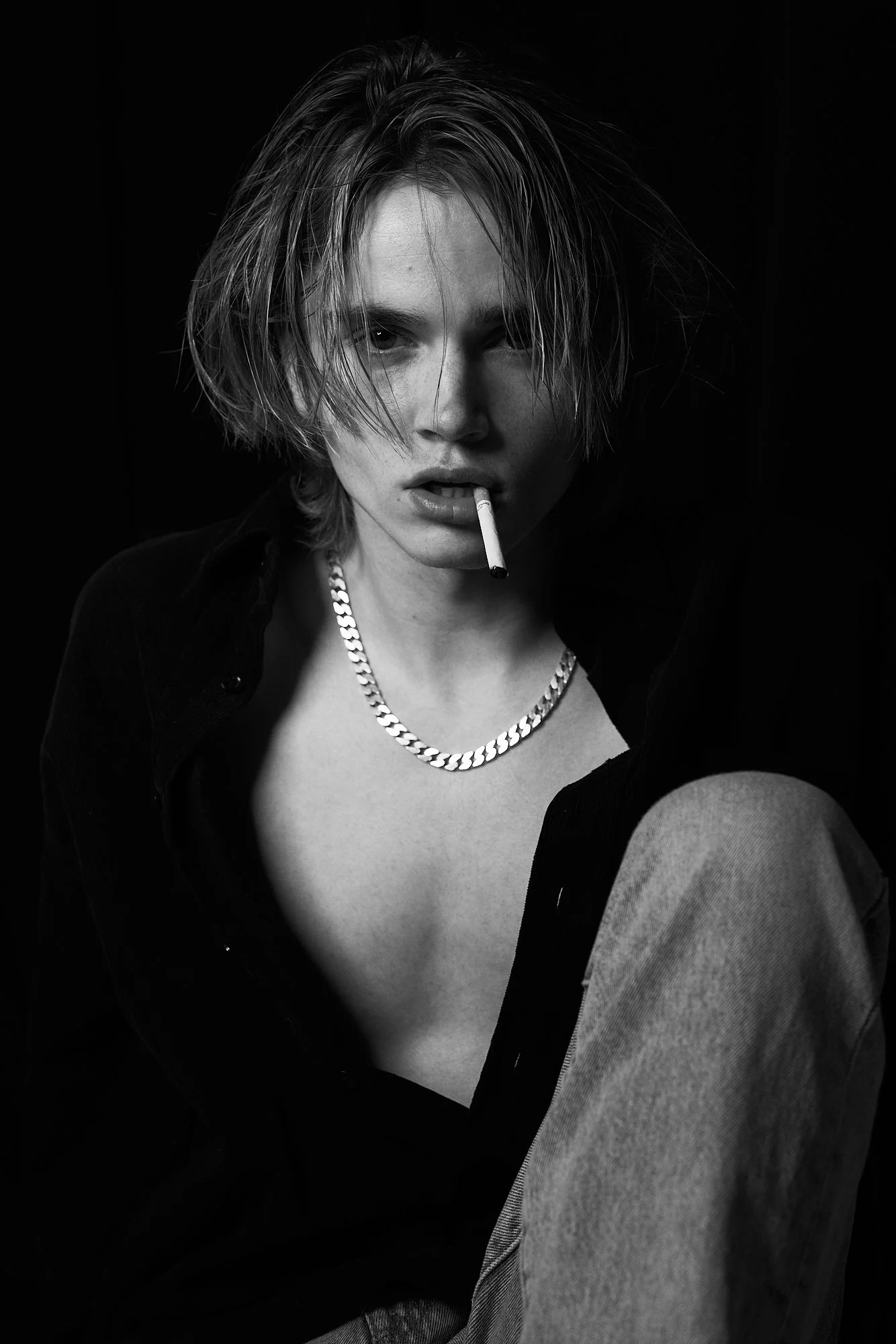 Black and white photo of a young woman with tousled hair, wearing a chain necklace, with a cigarette in her mouth, dressed in a dark shirt and light-colored pants.
