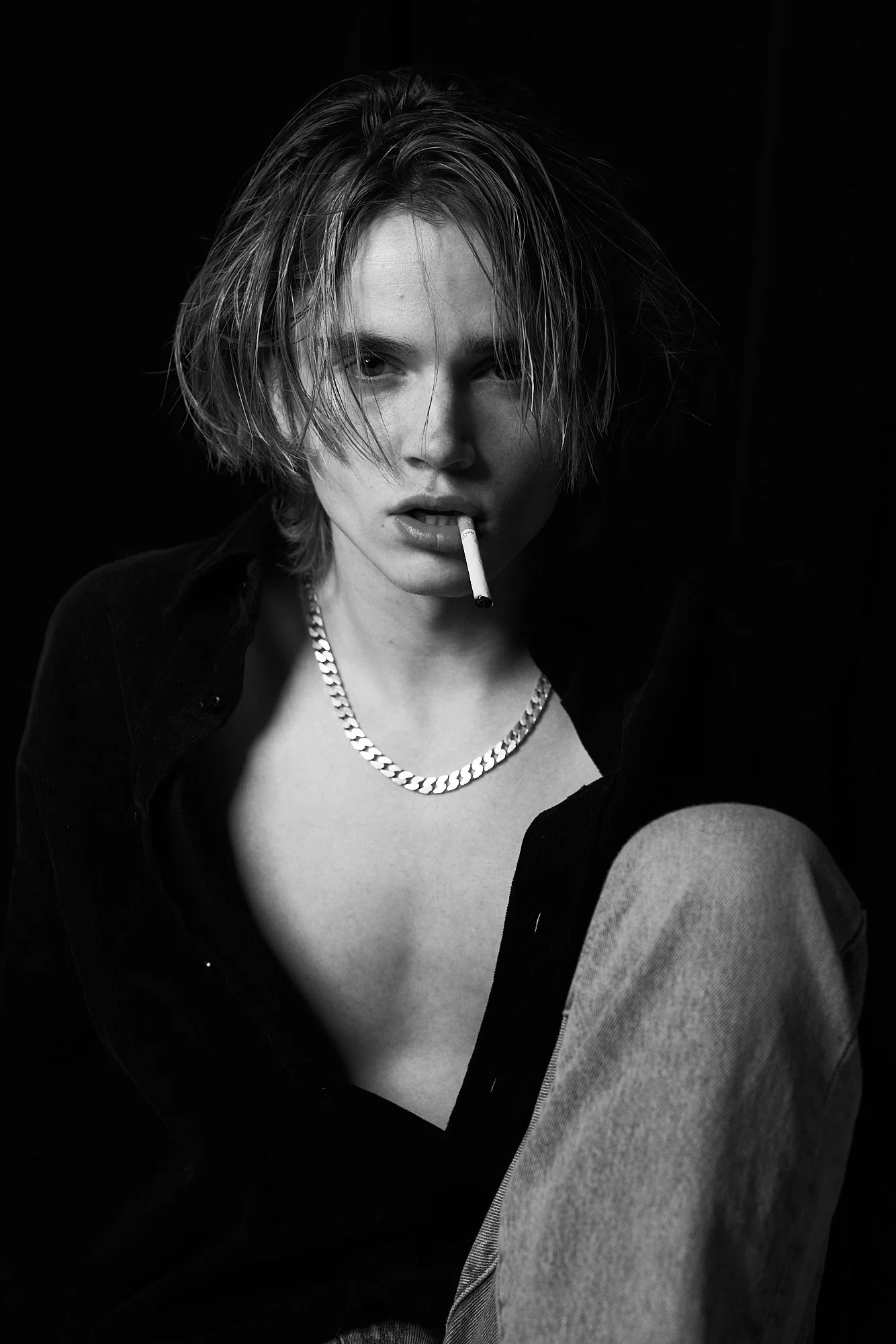 Black and white portrait of a woman with short, messy hair, wearing a chain necklace, a partially open shirt, and jeans, with a cigarette in her mouth.