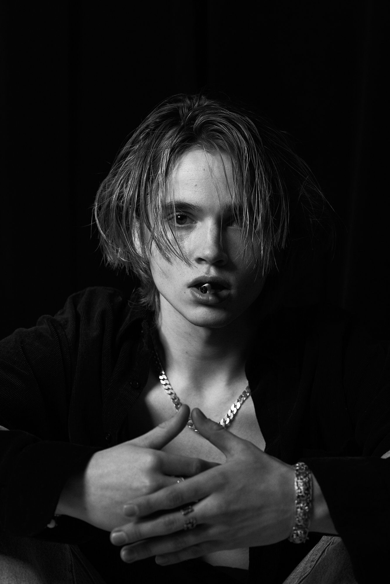 Black and white portrait of a young person with messy, shoulder-length hair, wearing a necklace, bracelet, and rings, with partially open lips and intense gaze.