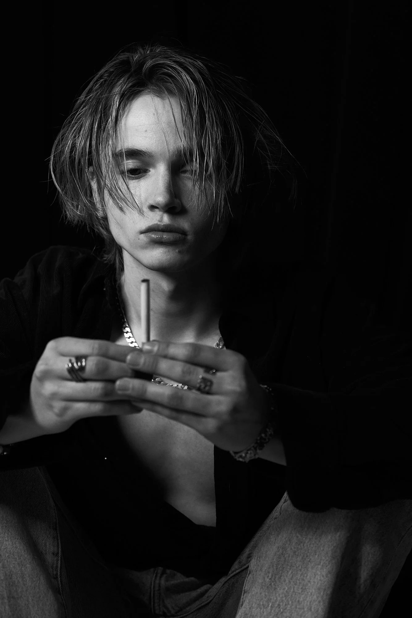 A woman with short, messy hair is sitting and looking down at her phone, holding a cigarette in her hands, in a black and white photo.