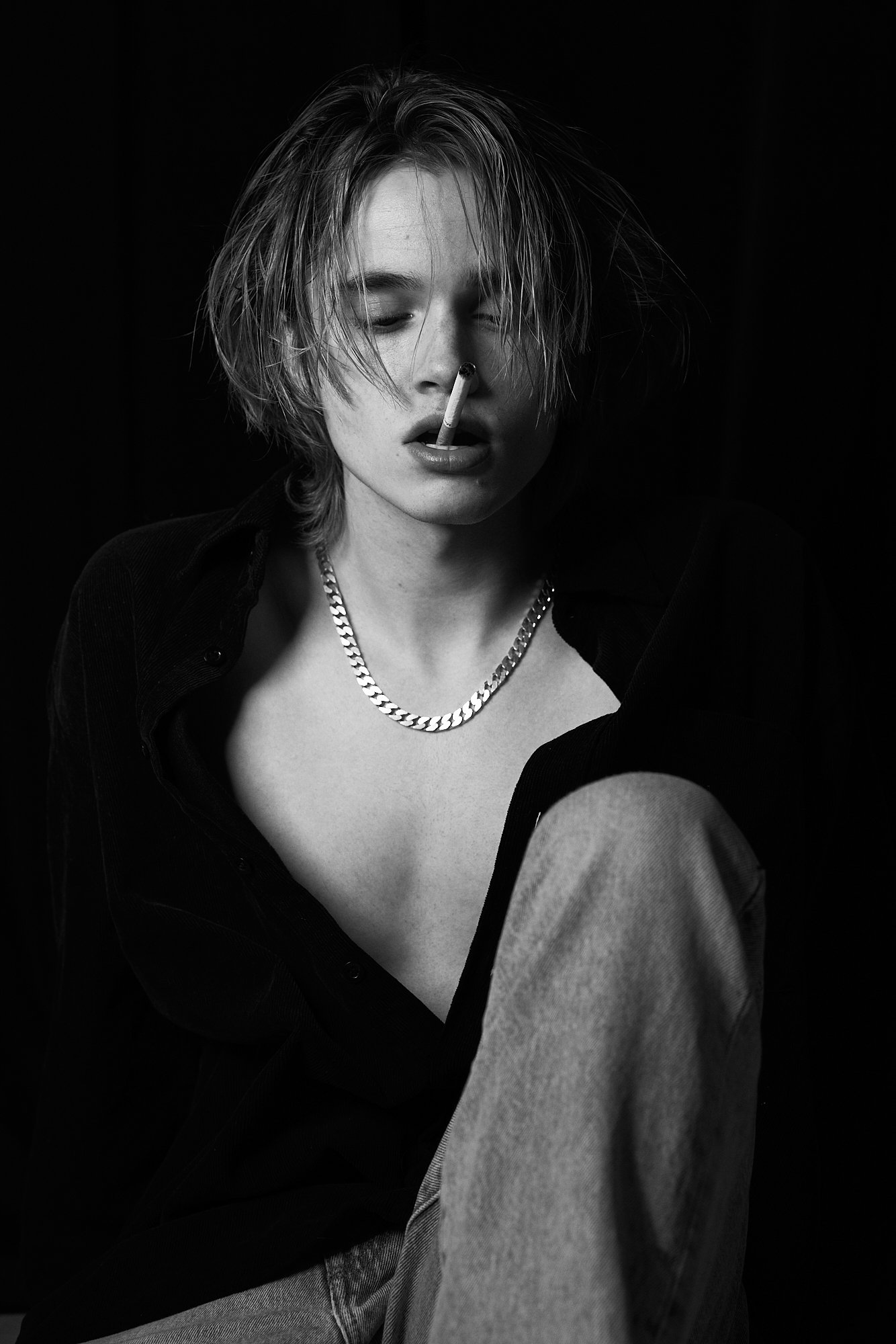 Black-and-white photograph of a young woman with short messy hair, wearing a chain necklace, an open shirt, and light-colored pants, with a cigarette in her nose.