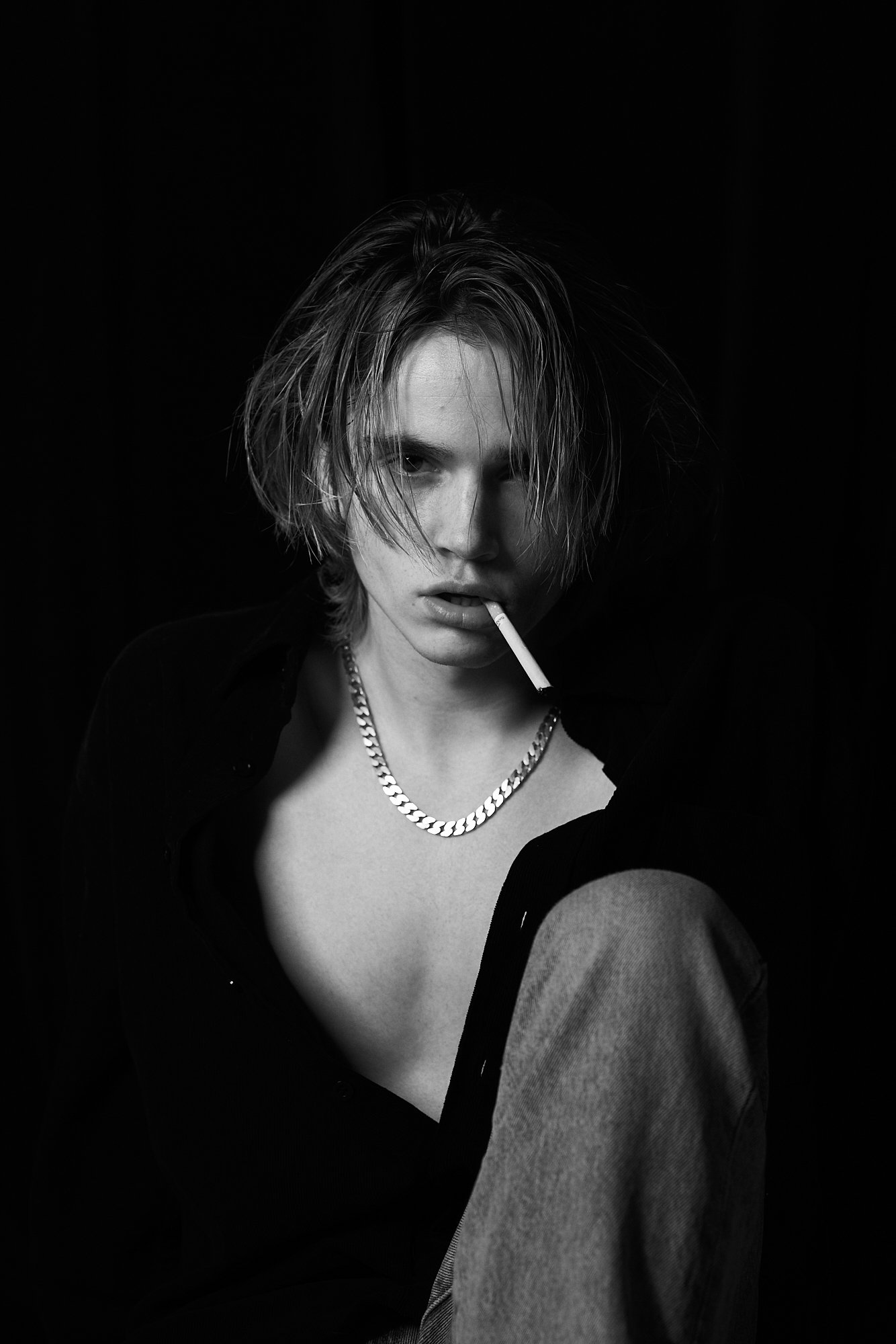 A black and white portrait of a young person with tousled hair, wearing a chain necklace, and holding a cigarette in their mouth, with a dark background.