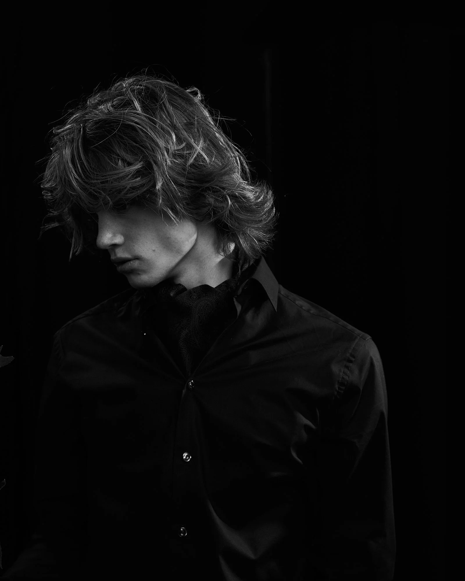 Black and white portrait of a person with wavy hair, wearing a button-up shirt and looking downward, against a dark background.