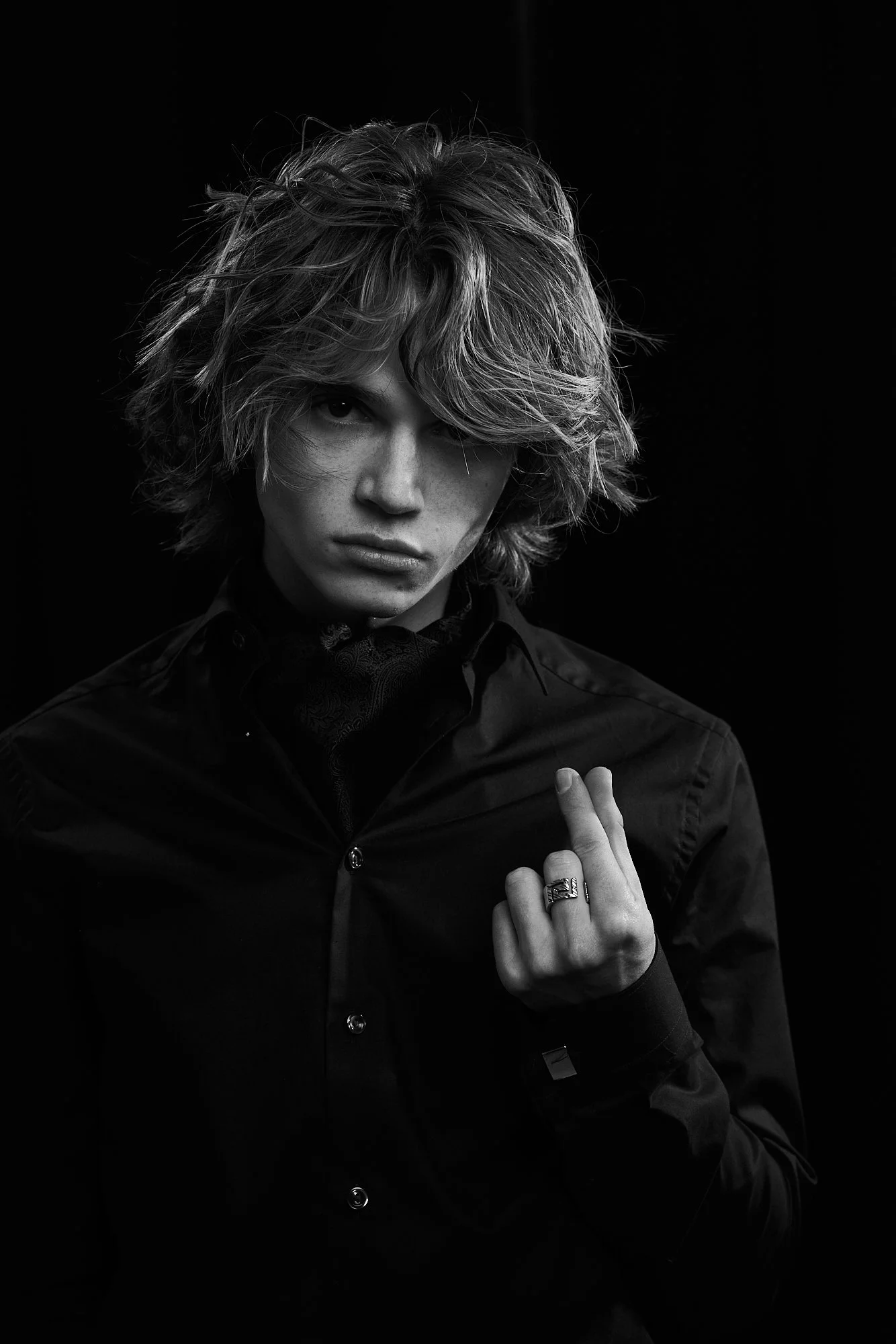 Black and white portrait of a young person with wavy, tousled hair, wearing a dark shirt and ring, looking directly at the camera with a serious expression, against a black background.