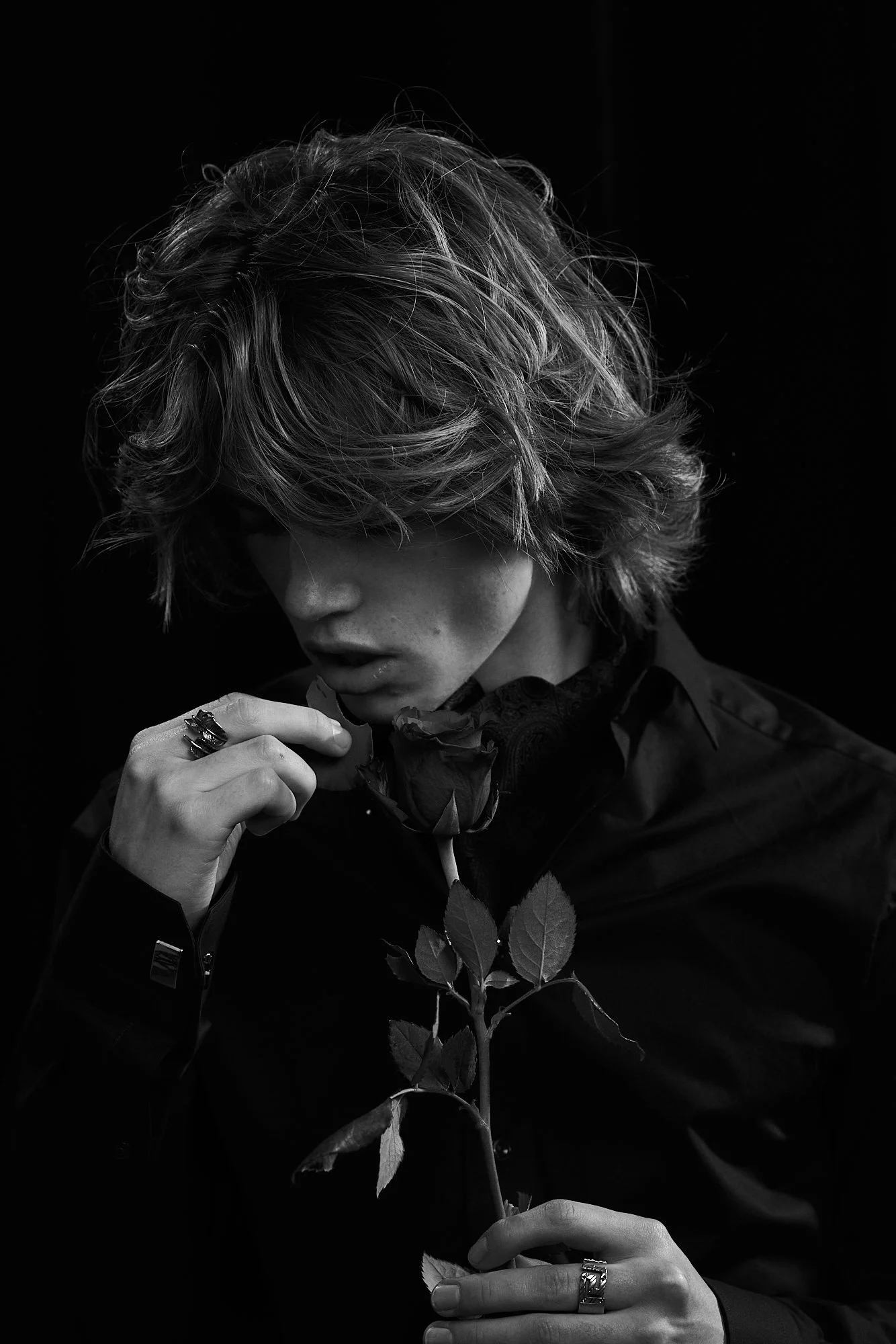 Black and white portrait of a person with wavy hair, holding a rose and looking down with a contemplative expression.
