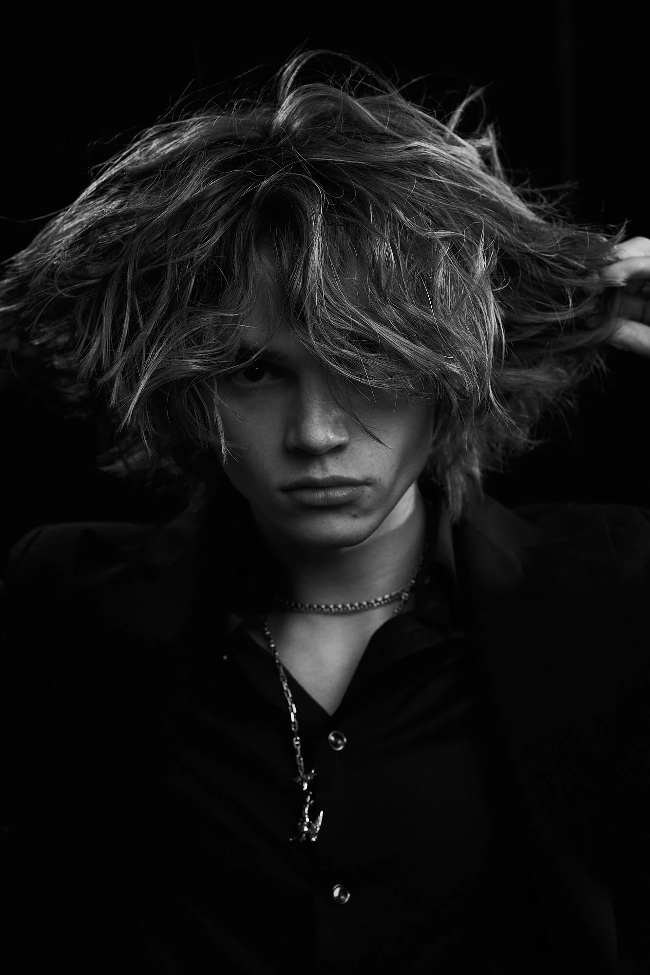 Black and white portrait of a person with messy, voluminous hair, looking directly at the camera with a serious expression, wearing a dark shirt and layered necklaces.