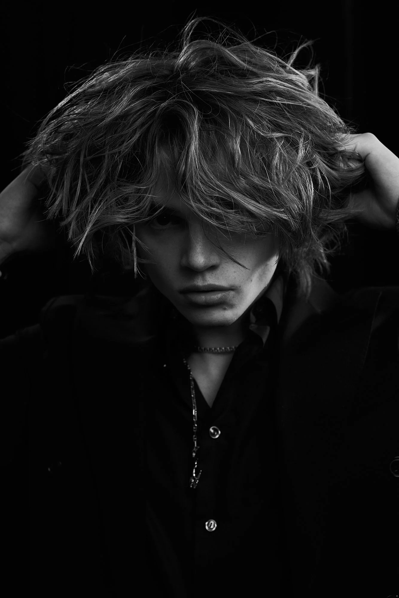 Black-and-white portrait of a young person with tousled, messy hair partially covering one eye, wearing a dark shirt and necklace, against a dark background.