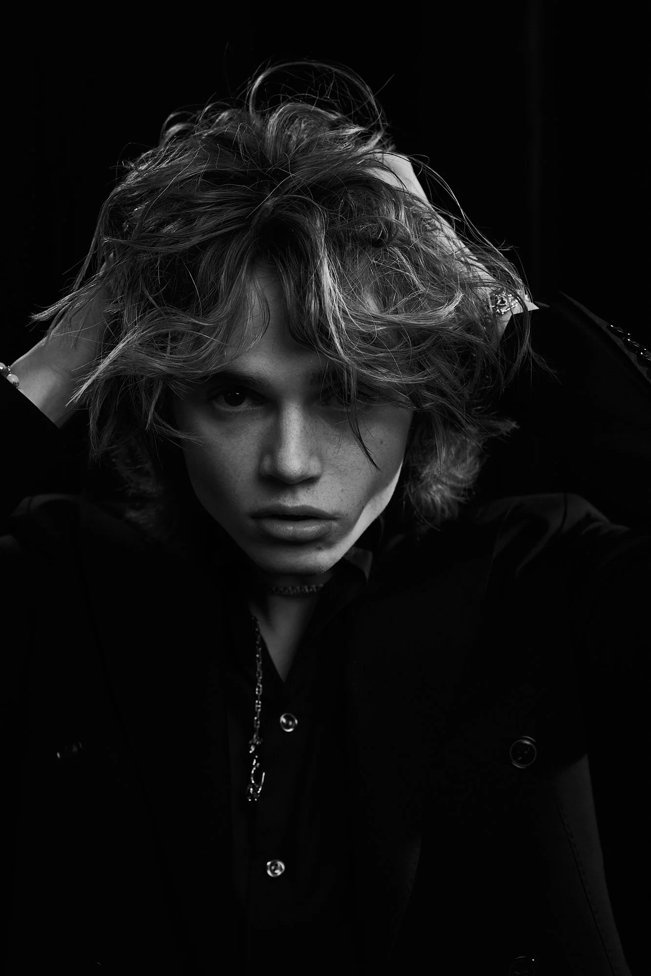 Black and white portrait of a woman with curly hair, intense gaze, dressed in dark clothing with jewelry, posing with hands on her head.