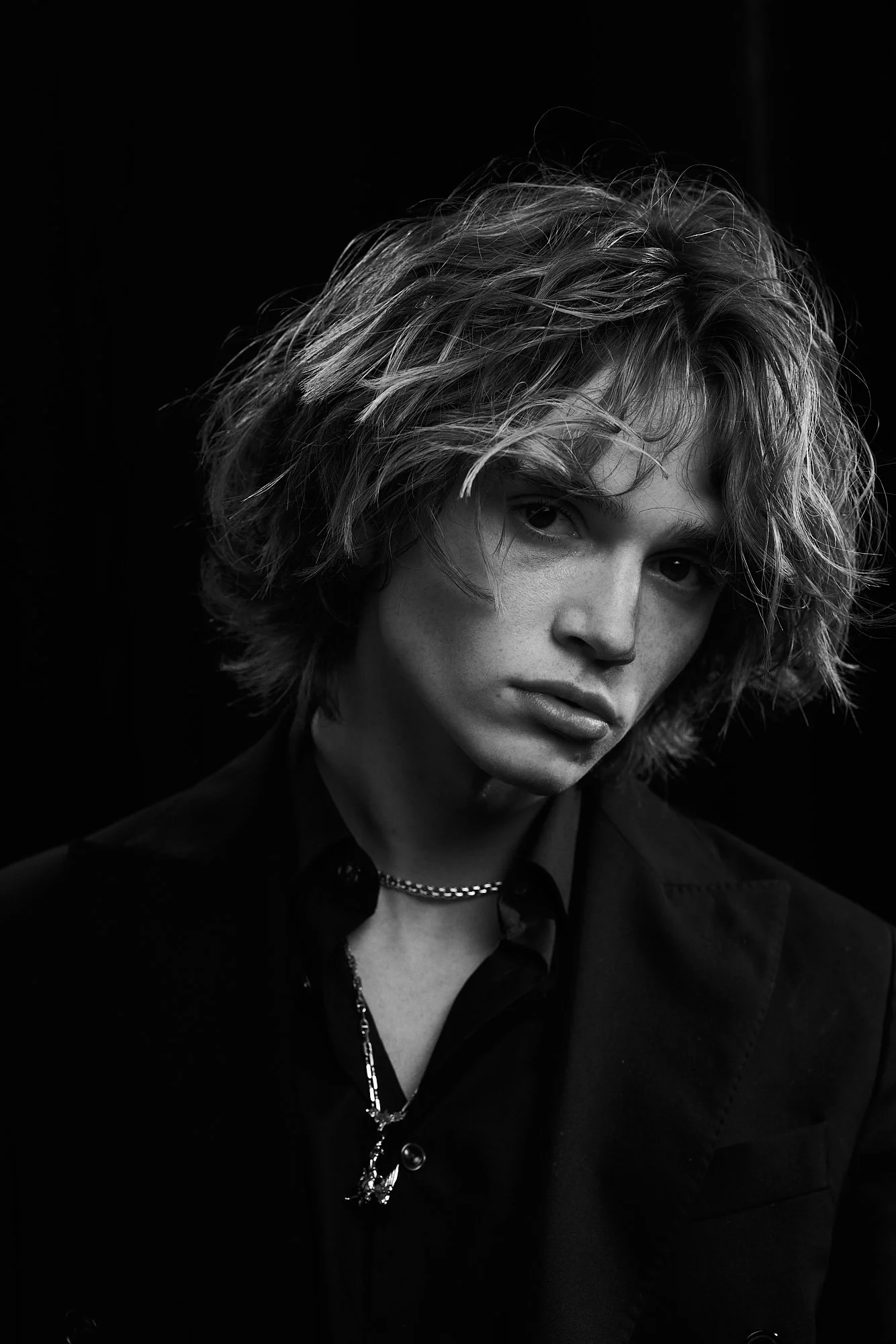 Black and white portrait of a young woman with tousled hair, wearing a dark blazer and layered necklaces, against a dark background.