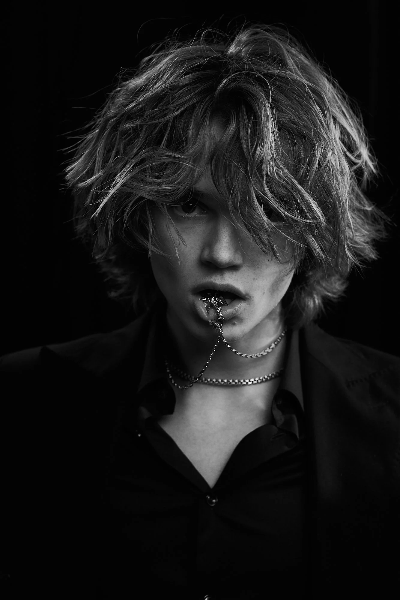 Black and white portrait of a person with tousled, shoulder-length hair, partially obscuring their face, biting a chain necklace, wearing a dark shirt and layered necklaces.