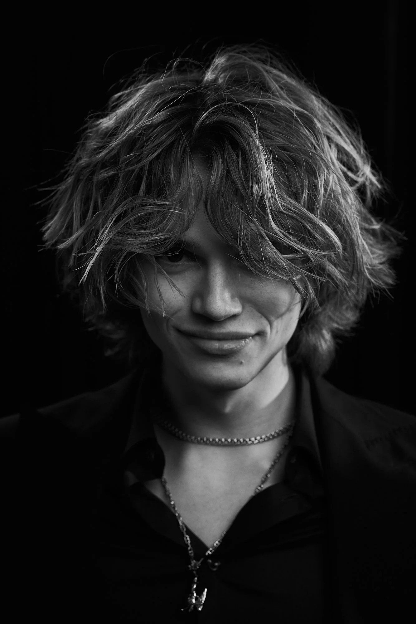 Black and white portrait of a woman with messy, shoulder-length hair, wearing a dark shirt and layered necklaces, smiling subtly at the camera.
