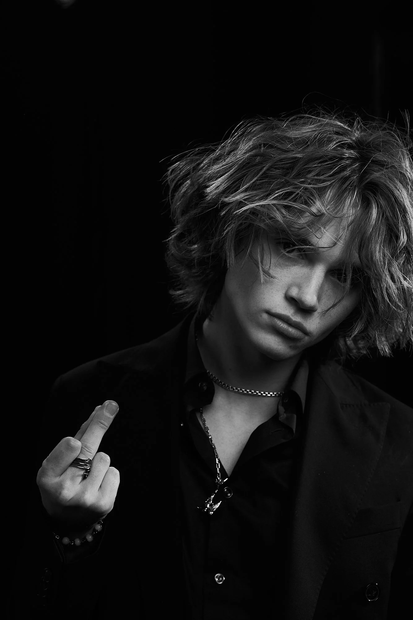 Black and white photo of a young man with tousled hair wearing a dark blazer, a chain necklace, rings, and bracelets, making a middle finger gesture.