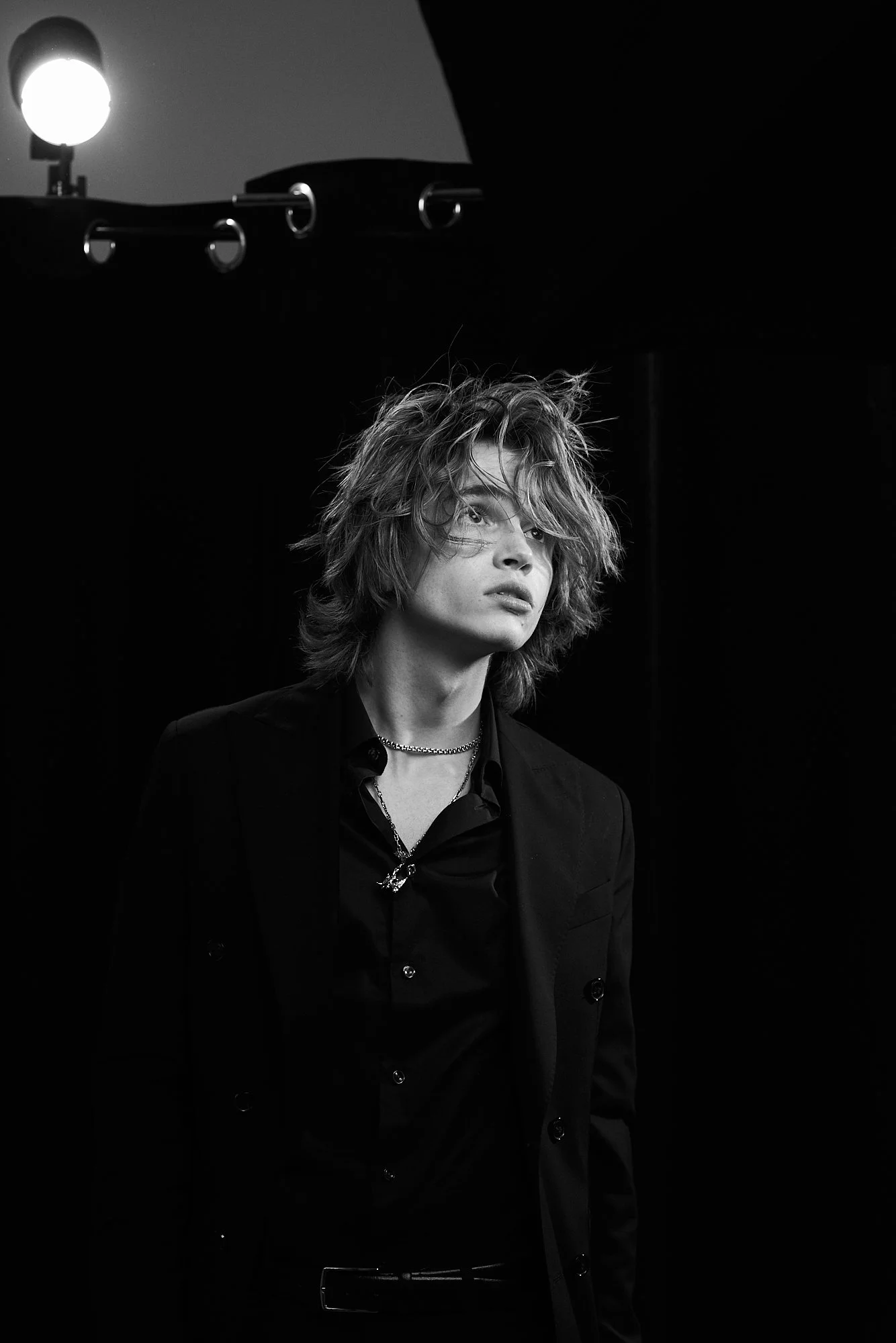 Black and white photo of a young man with messy hair, wearing a black blazer, black shirt, and layered necklaces, standing in a dimly lit room with a spotlight overhead.