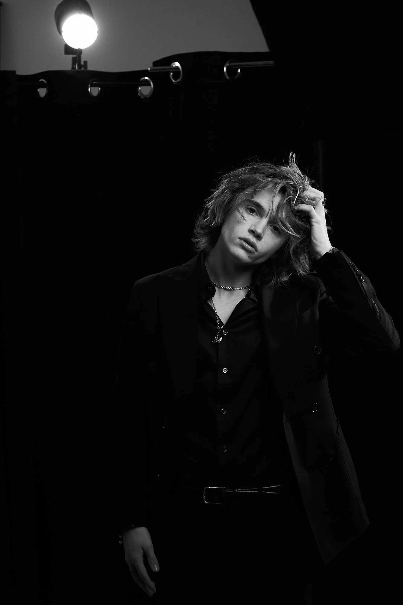 Black and white photo of a young person with casual, tousled hair, wearing dark clothing, standing in front of a dark background with a bright light source at the top left corner.