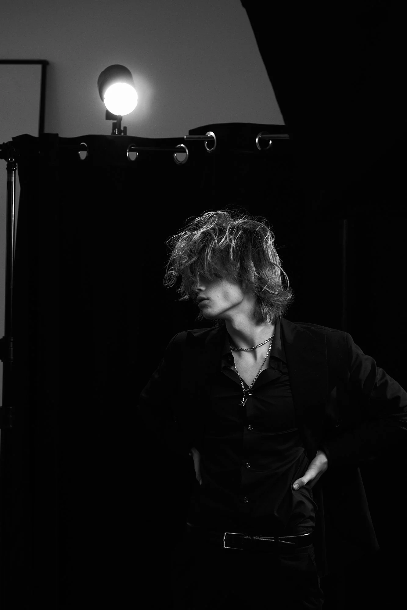 A person with wavy hair and layered hairstyle wearing a dark shirt and blazer, standing with hands on hips, in a dimly lit room with a black privacy screen and bright light in the background.