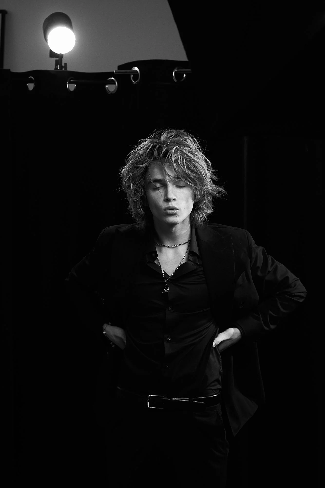 Black and white photo of a young man with messy hair, closed eyes, wearing a black shirt, blazer, and layered necklaces, standing in a dark room with a bright light source overhead.