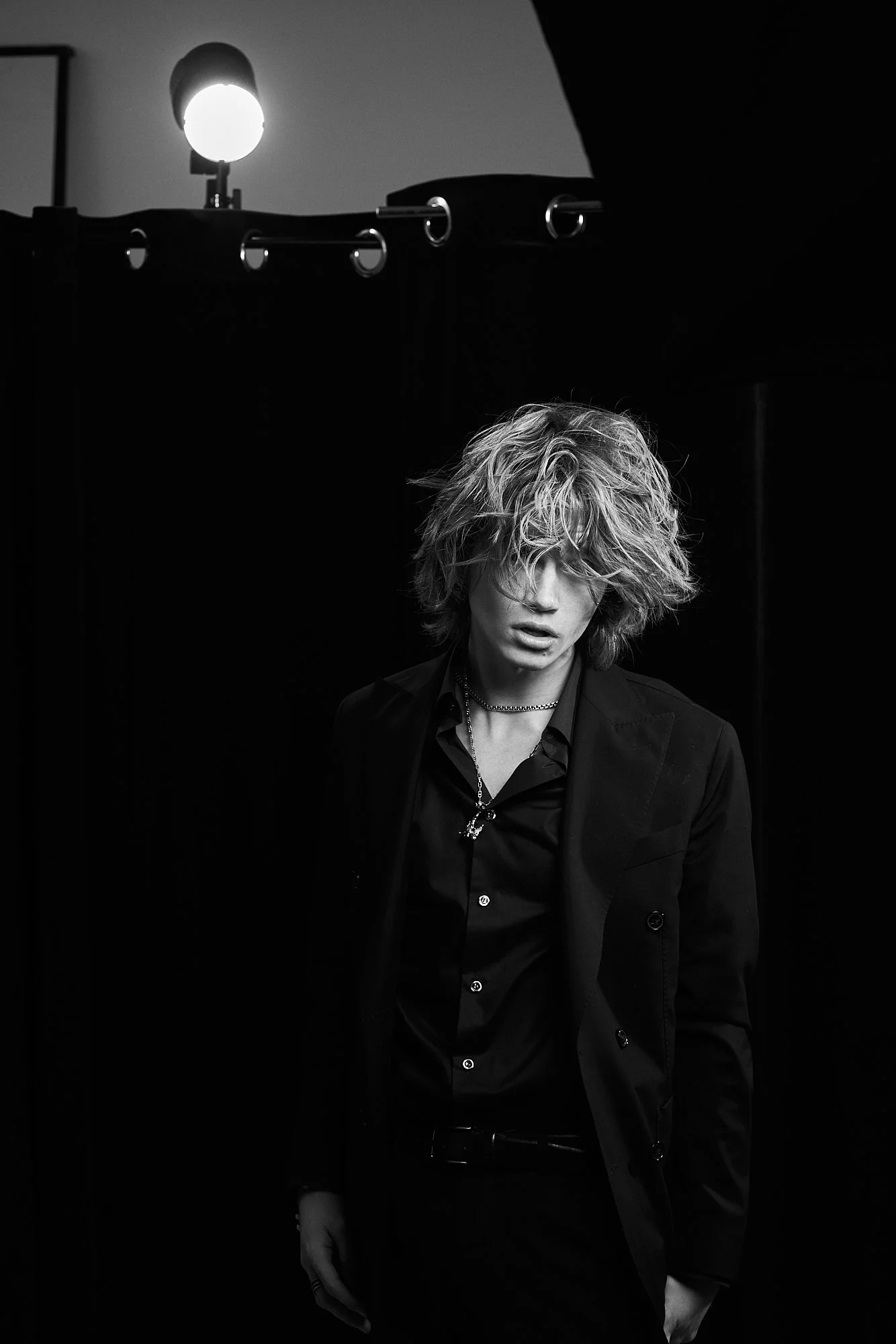A black and white photo of a person with curly, tousled hair, wearing a dark suit and jewelry, standing in front of a dark background with a bright light source behind them.