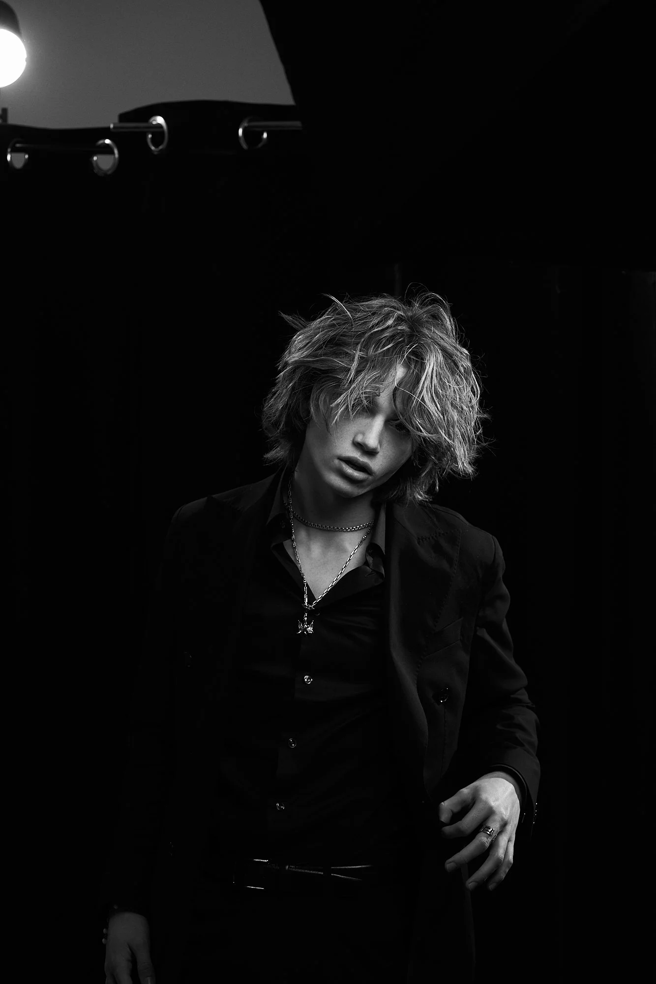 Black and white portrait of a young man with messy, shoulder-length hair wearing a dark blazer, open shirt, layered necklaces, and rings, posed against a dark background.