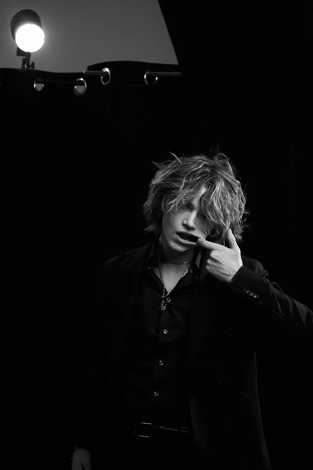 Black and white photo of a young man with messy hair, wearing a dark suit and shirt, making a playful expression with his finger touching his lips. A studio light is visible in the background.