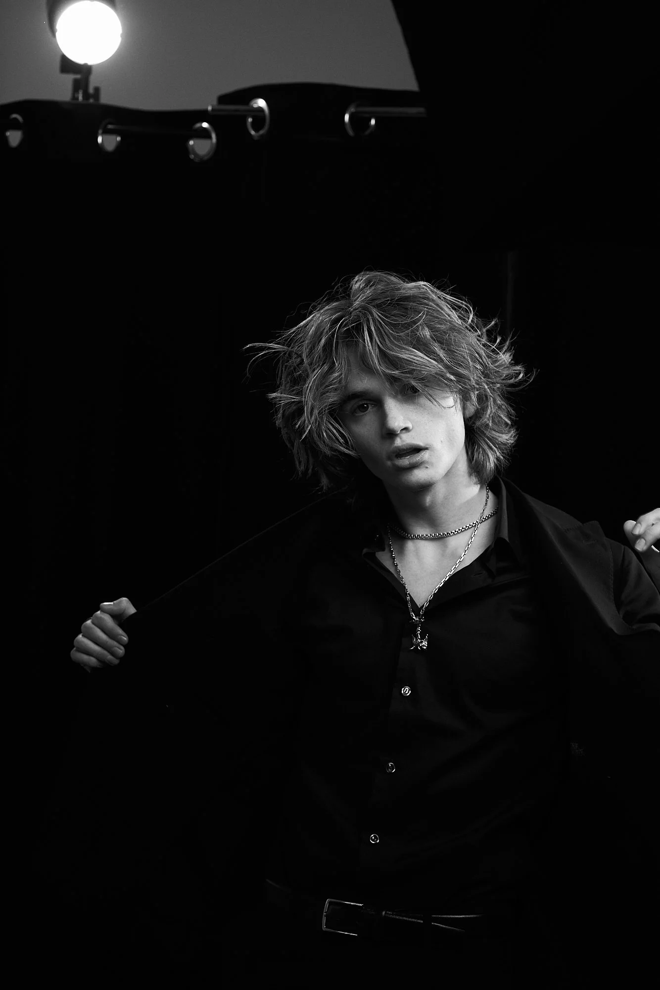 Black and white photo of a young man with tousled hair, wearing a black shirt and layered necklaces, posing against a dark background with studio lighting.