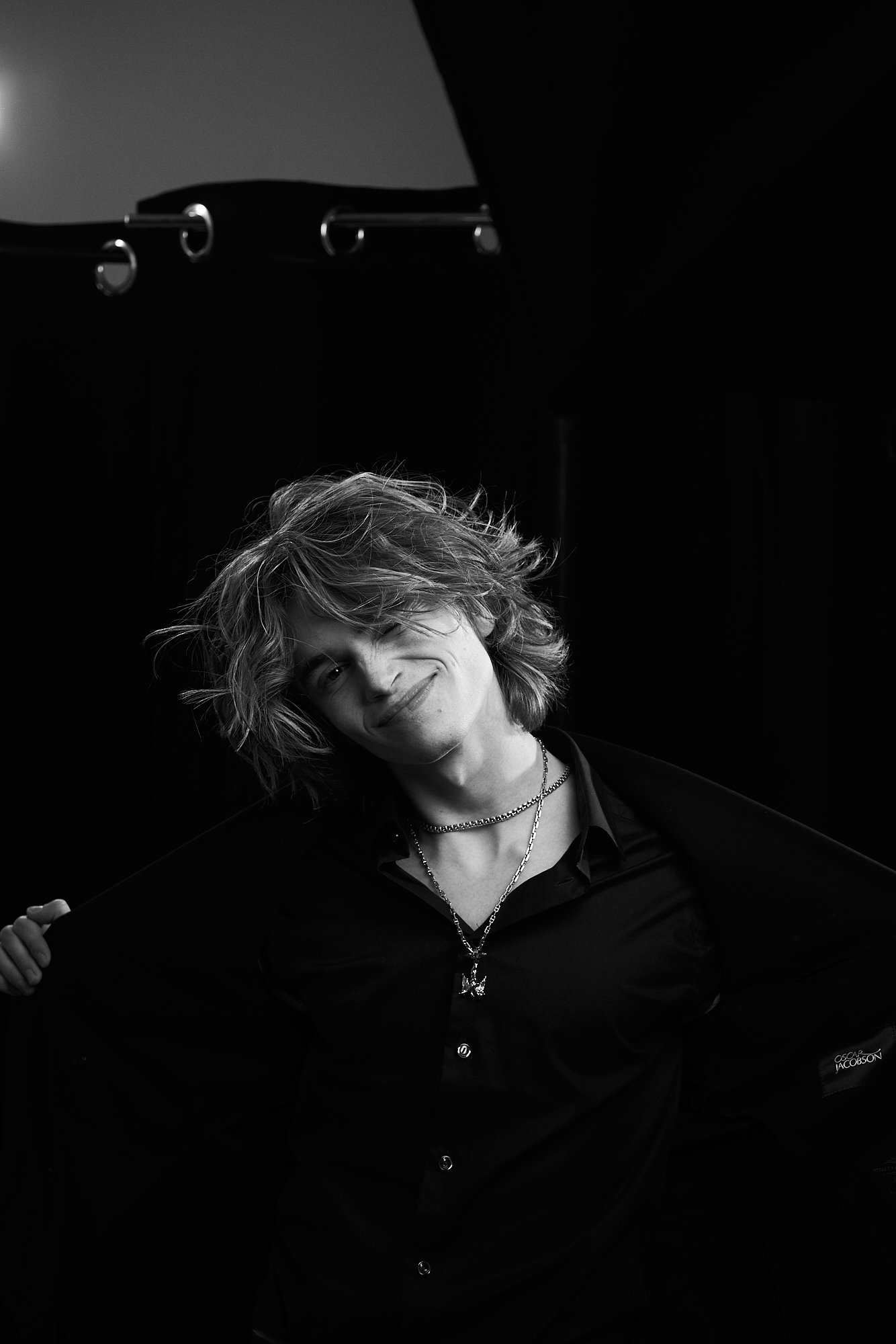 Black and white portrait of a young man with wavy, tousled hair, smiling and slightly tilting his head to the side, wearing layered necklaces and a dark shirt, in front of a dark background with metal curtain rings.