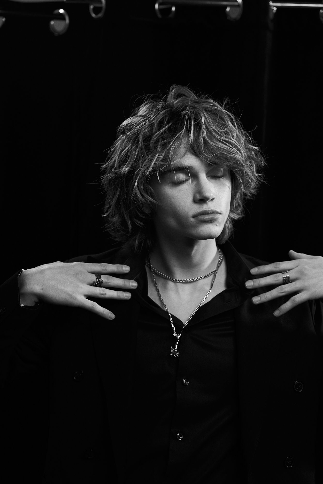 A black and white photo of a person with tousled hair, closed eyes, and androgynous appearance, wearing multiple necklaces, rings, and a dark jacket, with hands on their shoulders.