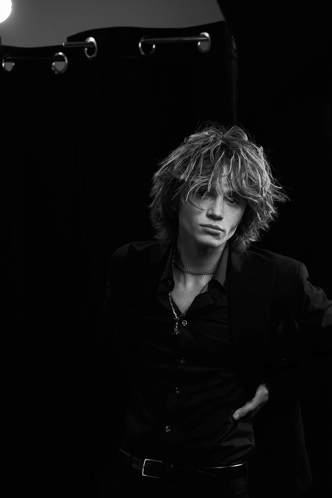 A young man with tousled, shoulder-length hair and light skin, wearing a black shirt and blazer, standing against a dark background with a curtain in the back.