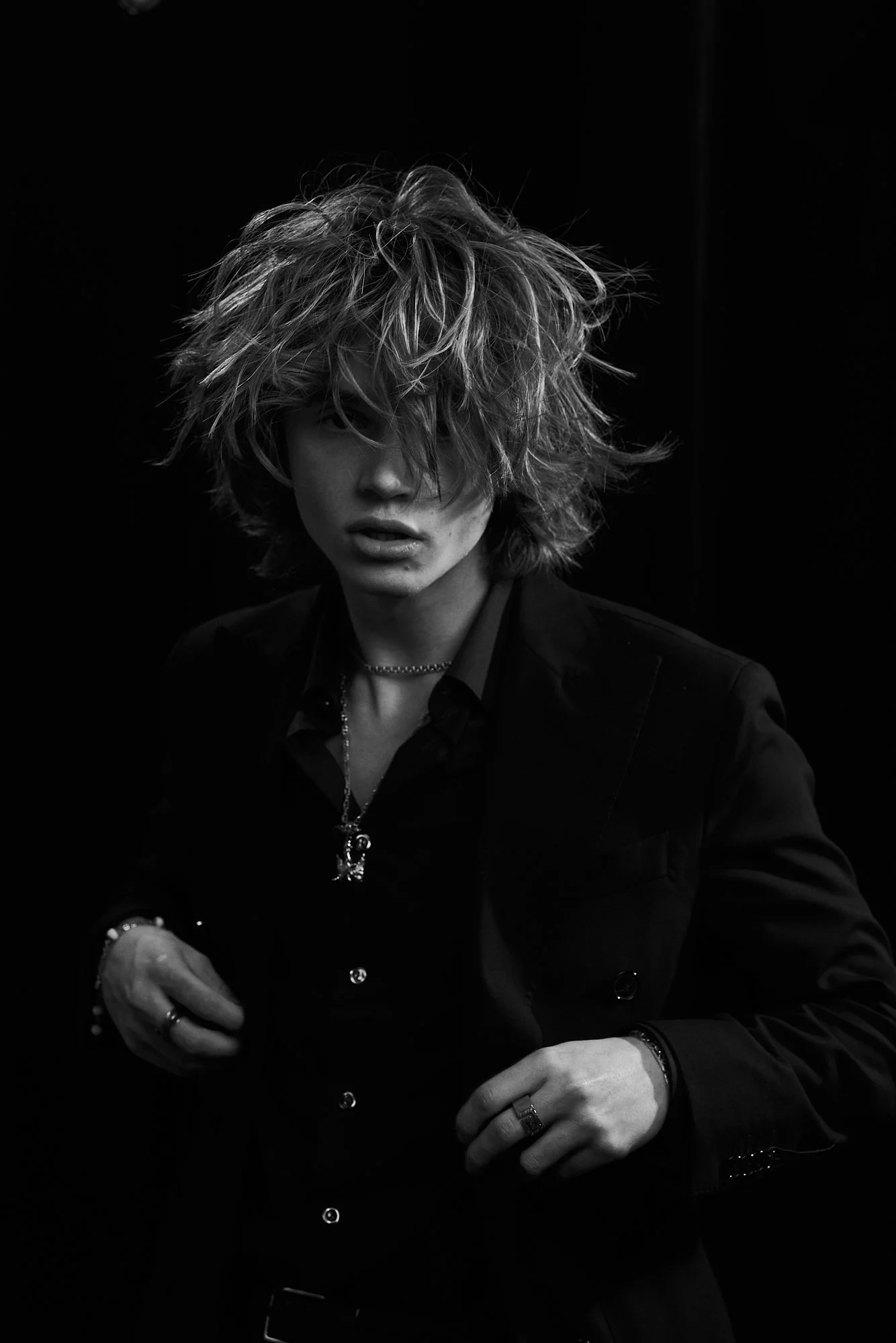 Black and white photo of a person with messy, curly hair, wearing a dark blazer, shirt, and jewelry, looking directly at the camera against a dark background.