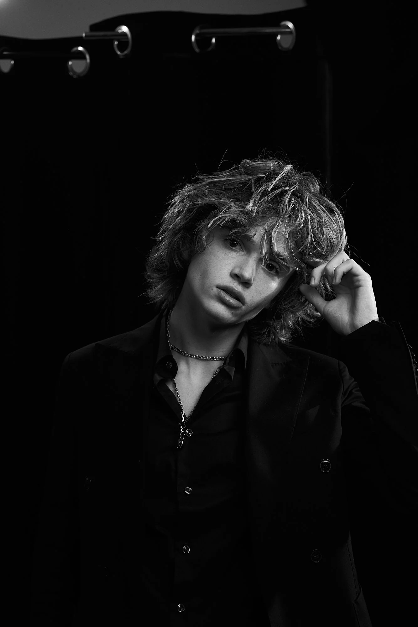 Black and white portrait of a young person with tousled hair, wearing a dark suit jacket and necklaces, with a dark background.